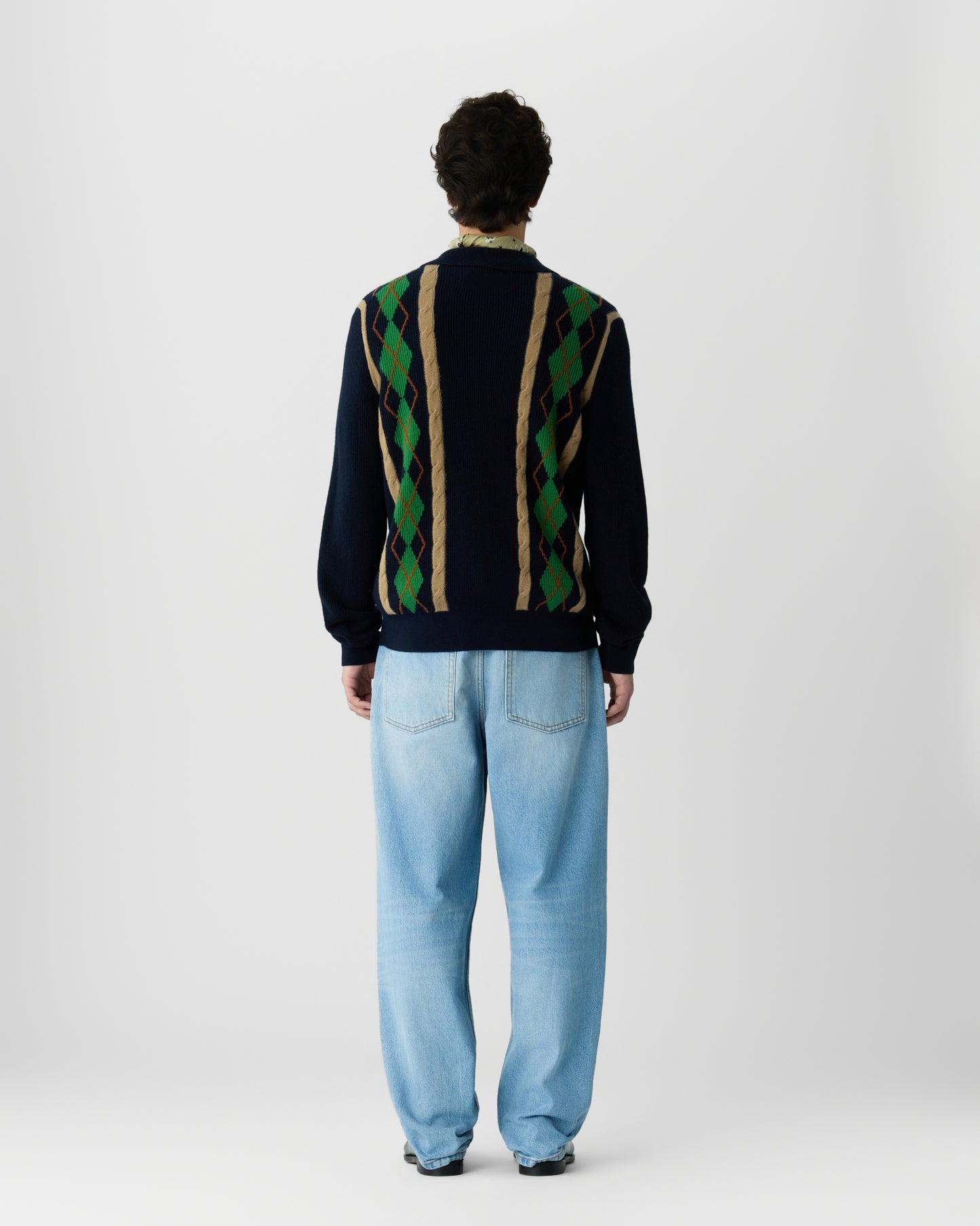 Man wearing a dark blue sweater with green, beige, and brown argyle pattern on the back and light blue jeans, standing facing away against a plain white background.