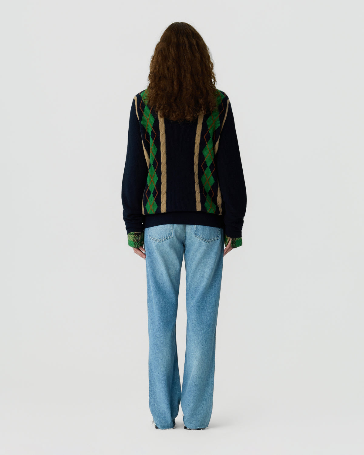 Back view of a woman wearing a dark cardigan with green and tan diamond stripes and light blue relaxed fit jeans against a plain white background