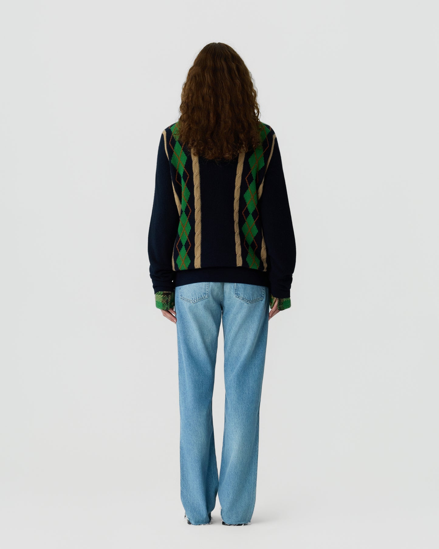 Person with long curly hair wearing a dark sweater with green and beige argyle and cable knit patterns, paired with light blue straight-leg jeans, standing facing away against a plain background.