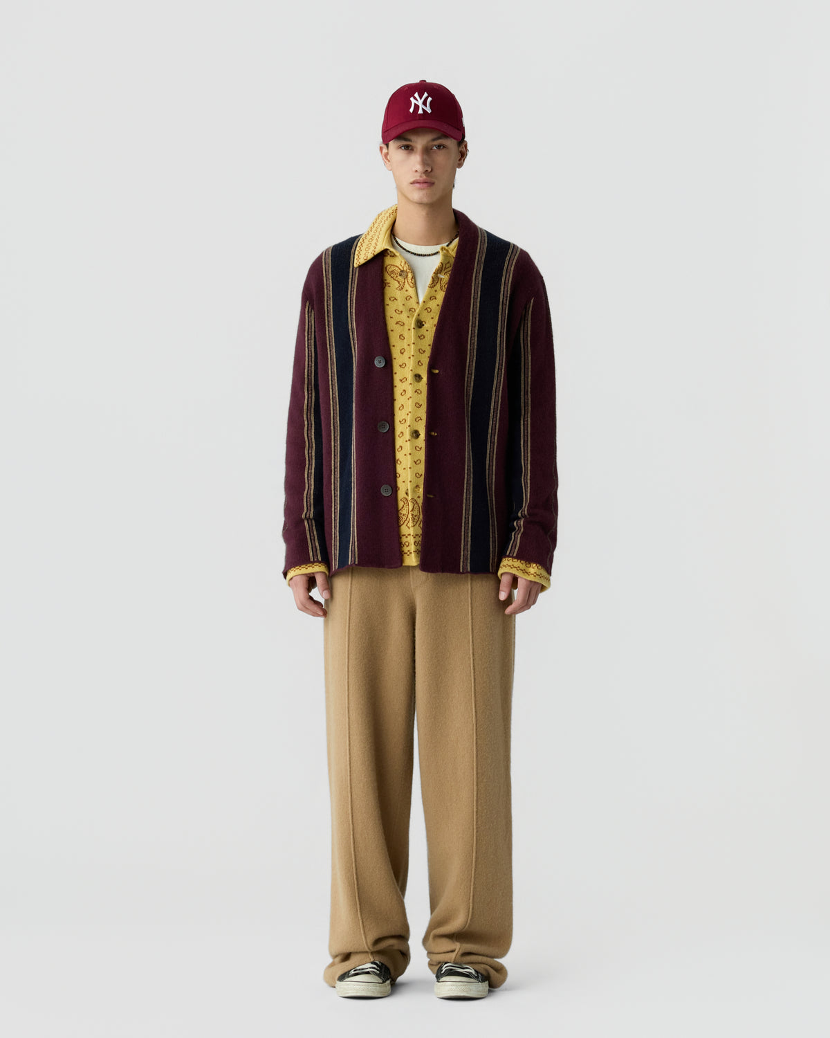 A young man wearing a red New York Yankees baseball cap, a striped maroon cardigan over a yellow patterned shirt, loose beige pants, and sneakers standing against a plain white background.