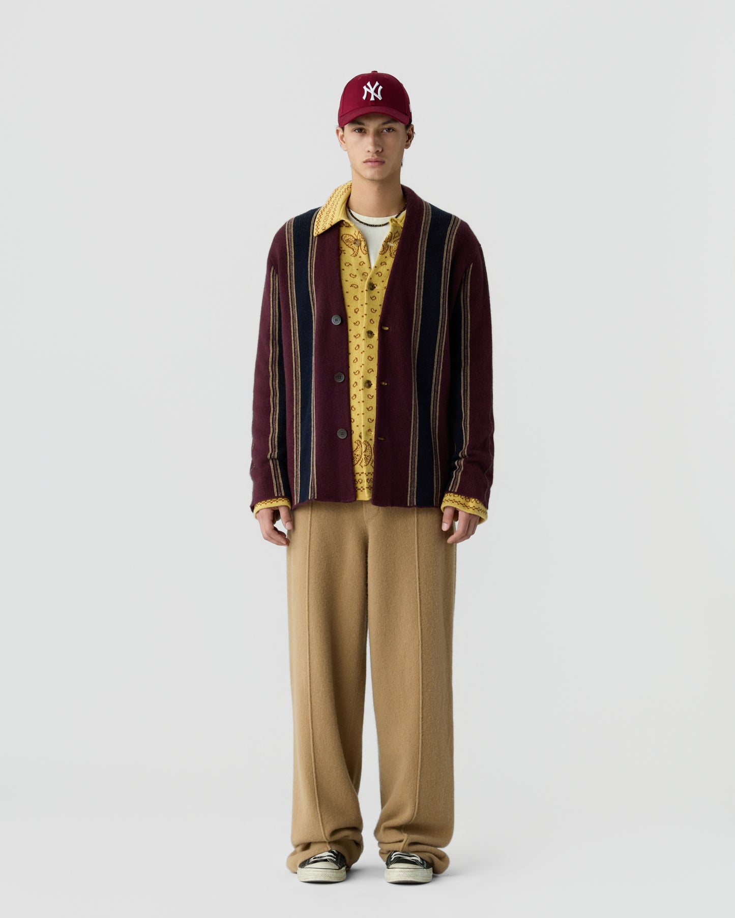A young man wearing a red New York Yankees baseball cap, a striped maroon cardigan over a yellow patterned shirt, loose beige pants, and sneakers standing against a plain white background.