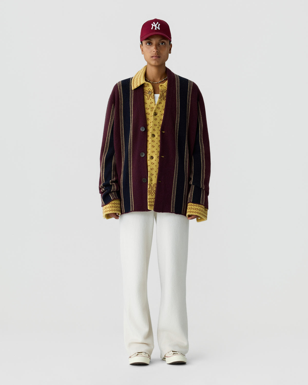 Person standing against a light gray background wearing a burgundy striped cardigan, patterned yellow shirt, white trousers, cream shoes, and a burgundy baseball cap