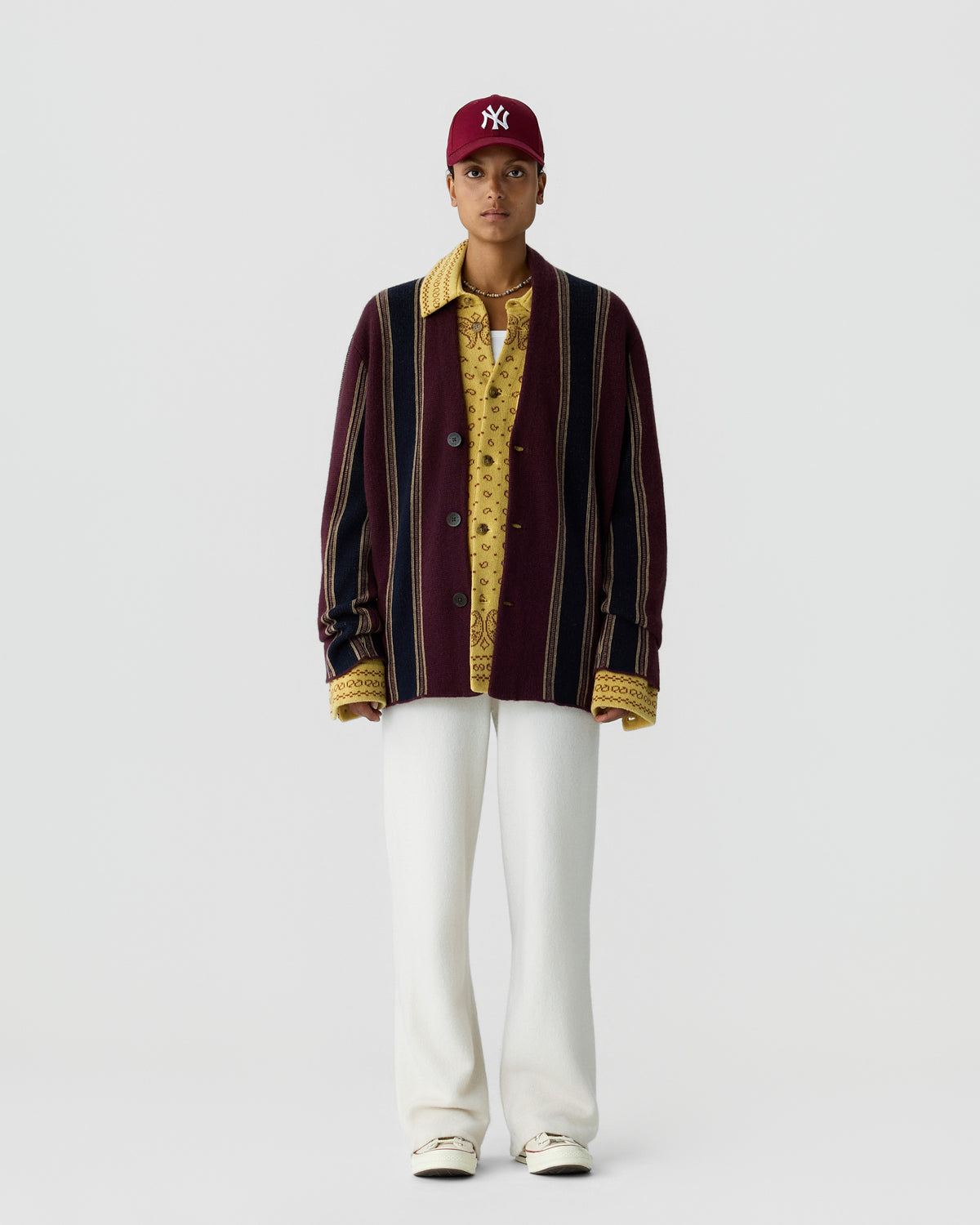 Person wearing a maroon and navy striped cardigan over a yellow patterned shirt, white pants, white shoes, and a maroon baseball cap with a white logo, standing against a plain light background.