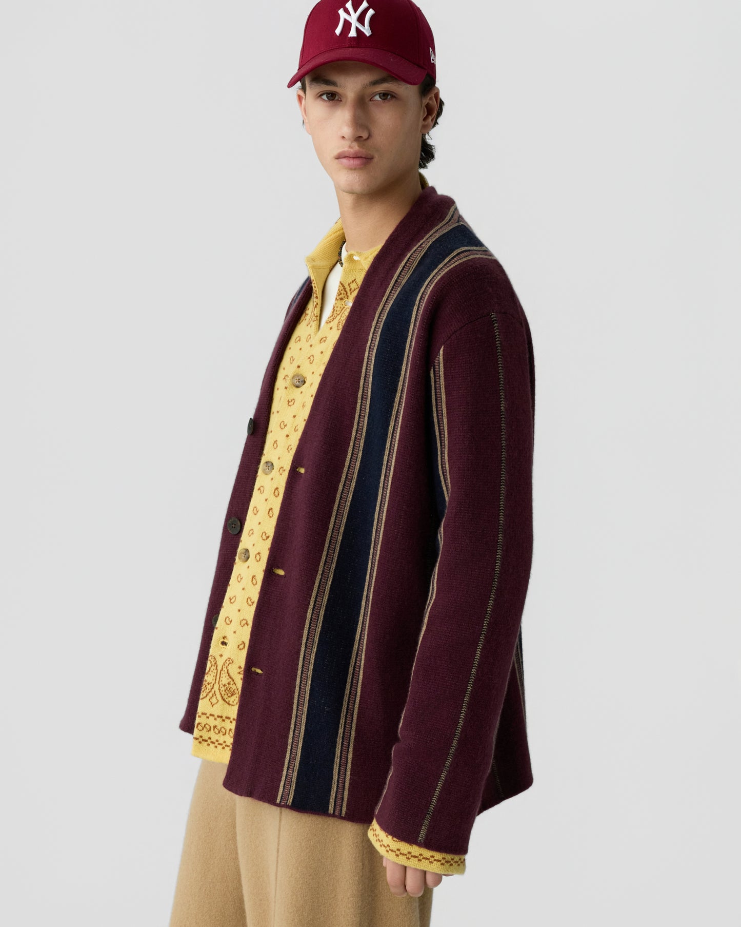 A young man wearing a maroon baseball cap and a maroon cardigan with vertical stripes over a yellow patterned shirt and beige pants, standing against a plain light gray background.