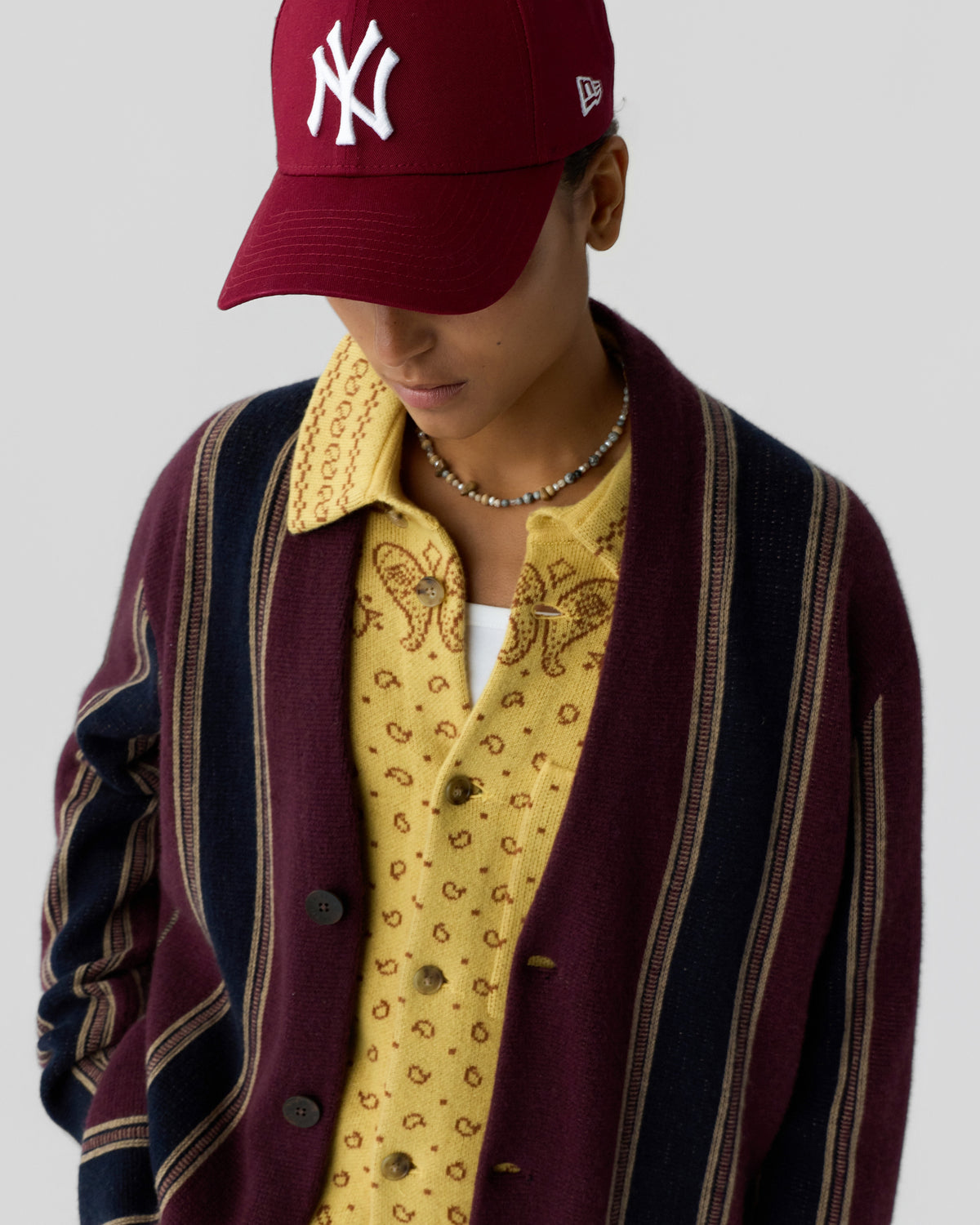 Person wearing a maroon and navy striped cardigan over a yellow patterned shirt with a white undershirt, paired with a maroon baseball cap featuring a white logo.