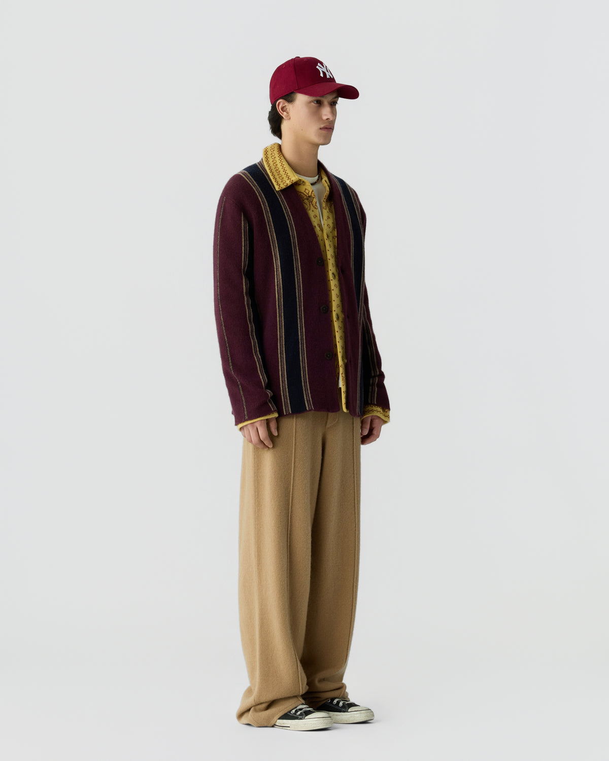 Side view of a young man wearing a burgundy striped cashmere jacket over a yellow patterned shirt, loose beige trousers, black and white sneakers, and a red baseball cap.