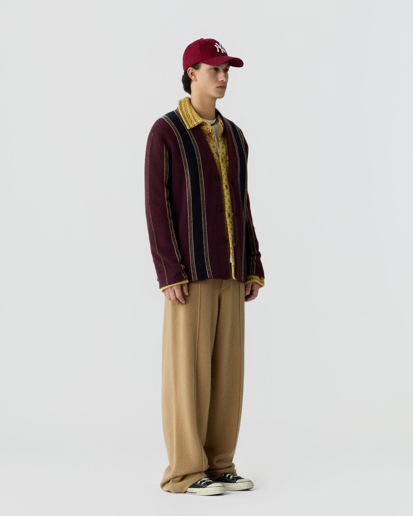 Side view of a young man wearing a burgundy striped cashmere jacket over a yellow patterned shirt, loose beige trousers, black and white sneakers, and a red baseball cap.