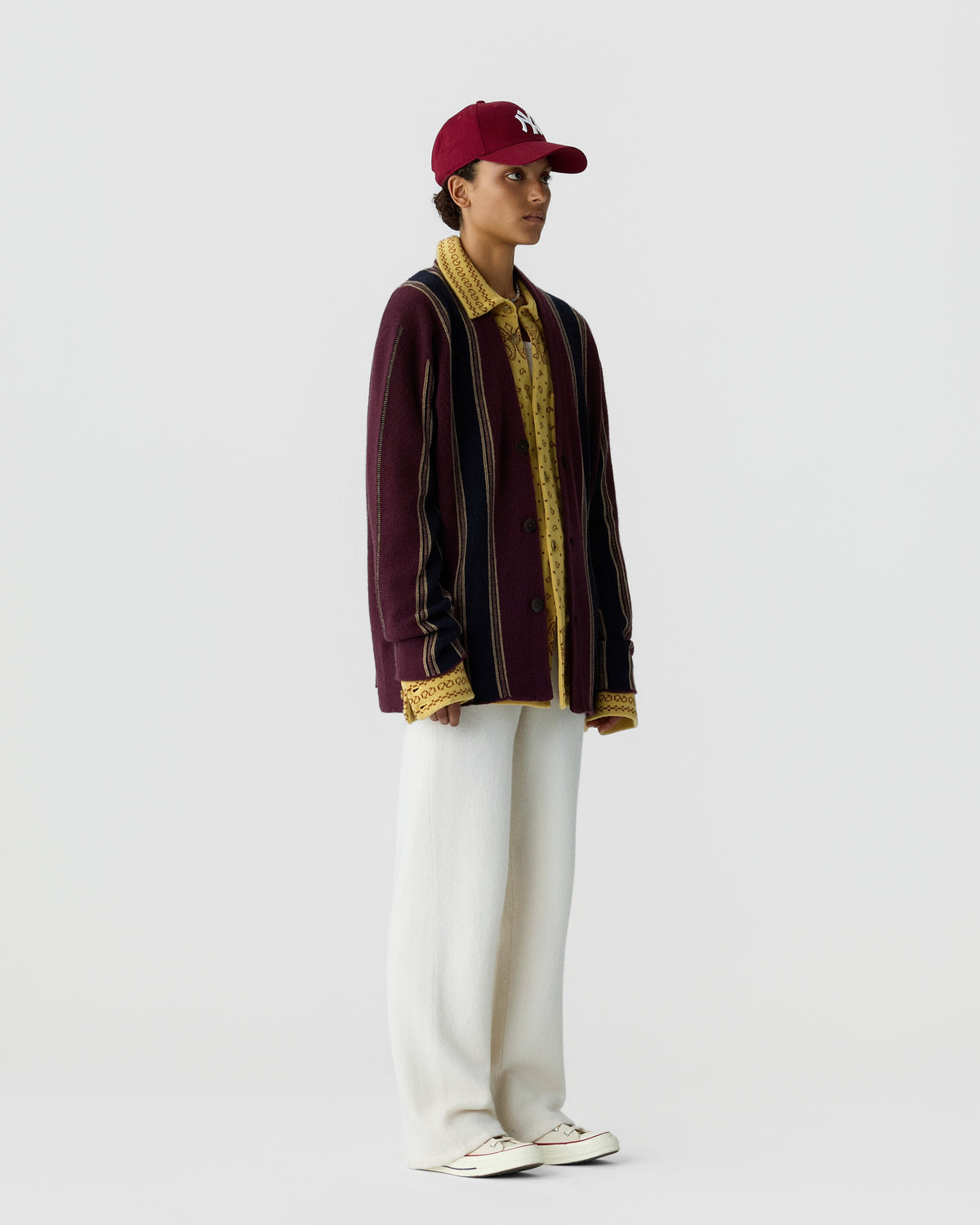 Young person wearing a maroon baseball cap, a brown and yellow knit cardigan with vertical stripes over a yellow patterned shirt, and loose white pants with white sneakers.