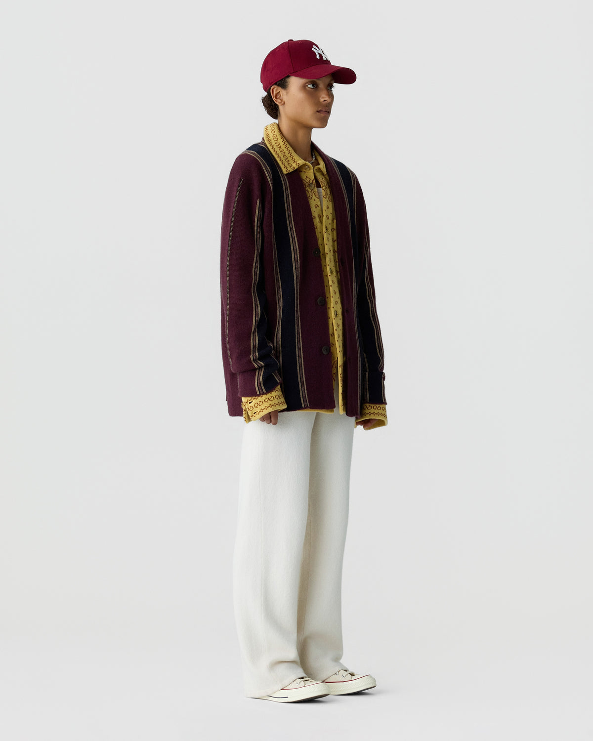 Model wearing a burgundy striped cardigan over a patterned shirt, white trousers, white sneakers, and a matching burgundy baseball cap against a light background