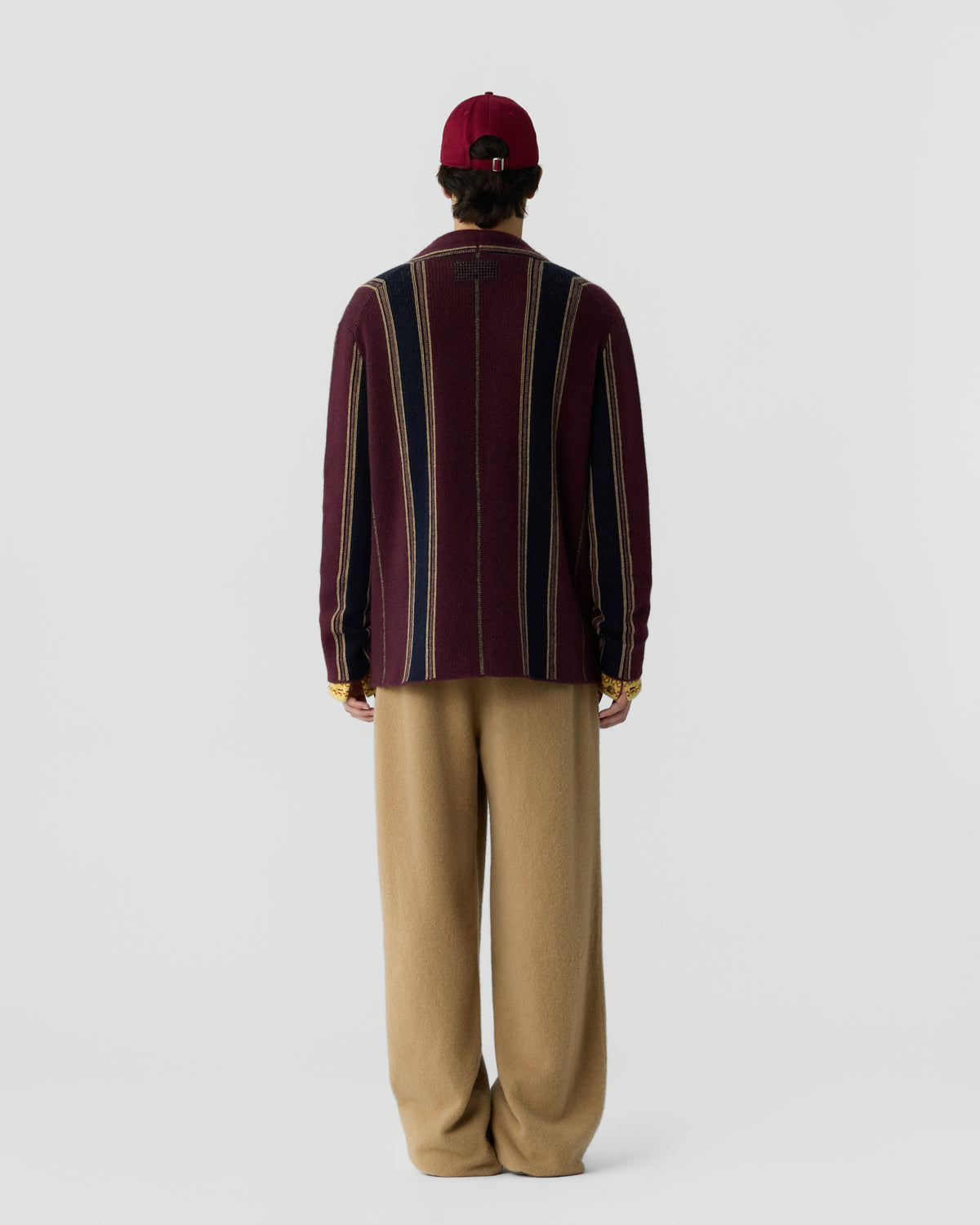 Person wearing a burgundy cap, a long-sleeve sweater with vertical stripes in burgundy, navy blue, and beige, paired with loose tan pants, standing facing away against a plain background.