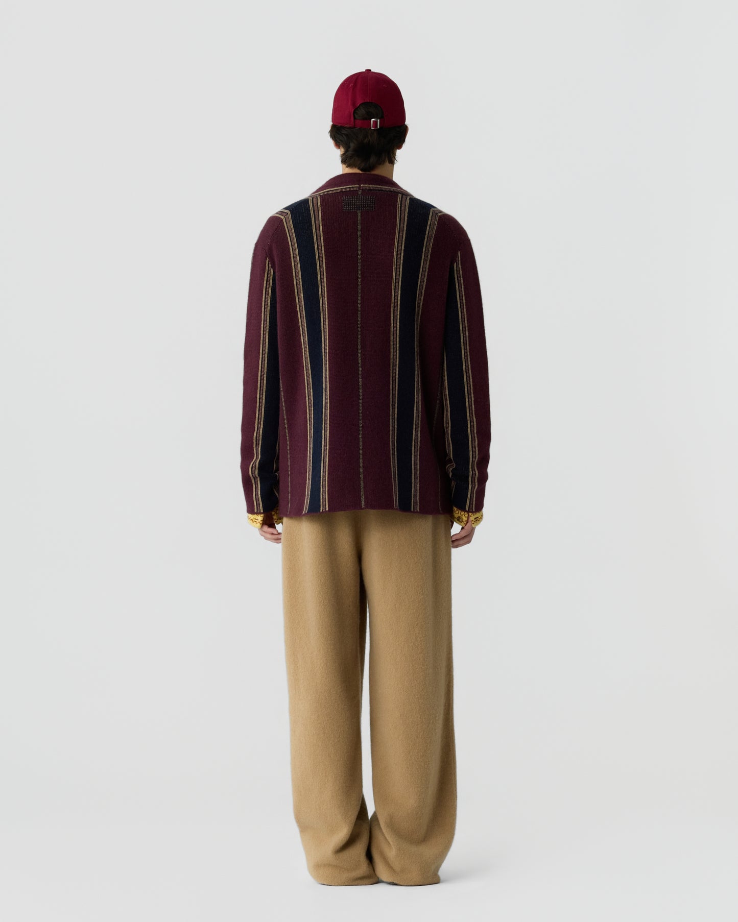 Person wearing a burgundy cap, a long-sleeve sweater with vertical stripes in burgundy, navy blue, and beige, paired with loose tan pants, standing facing away against a plain background.