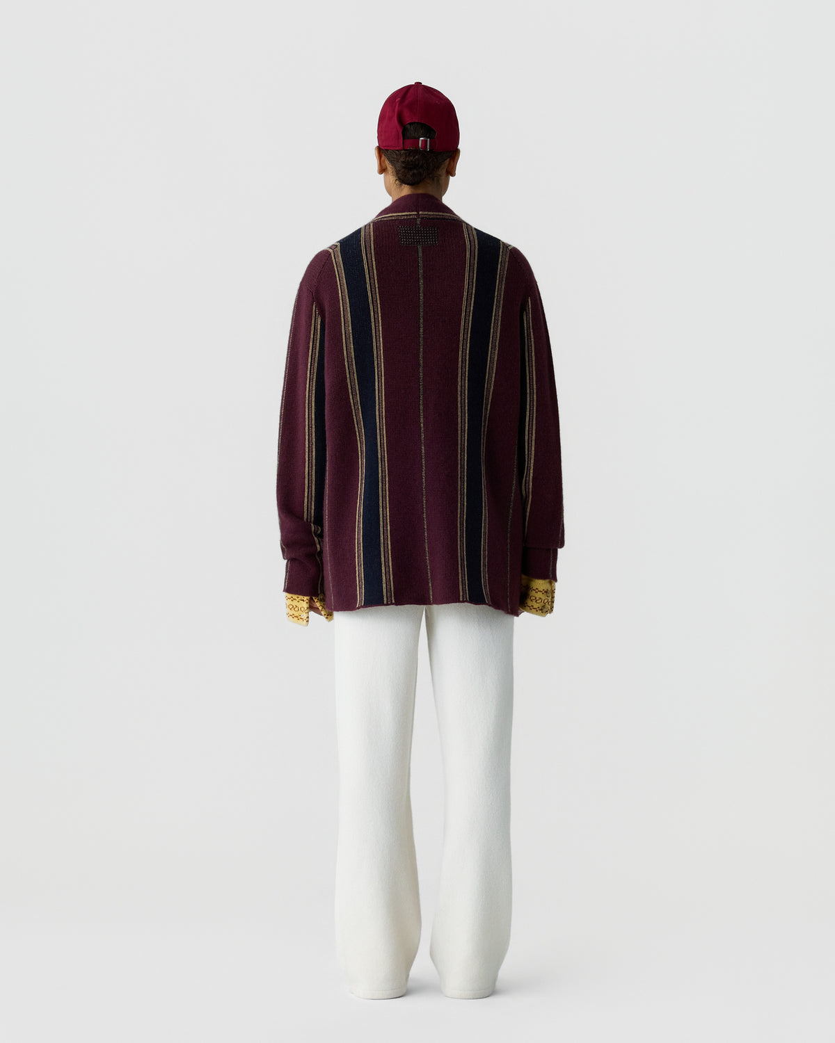 Person seen from behind wearing a striped long-sleeve sweater in burgundy, navy, and beige, white wide-leg pants, a red cap, and yellow gloves against a plain background.