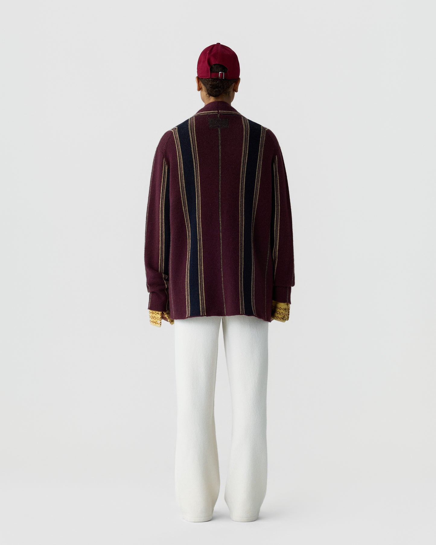 Person seen from behind wearing a striped long-sleeve sweater in burgundy, navy, and beige, white wide-leg pants, a red cap, and yellow gloves against a plain background.