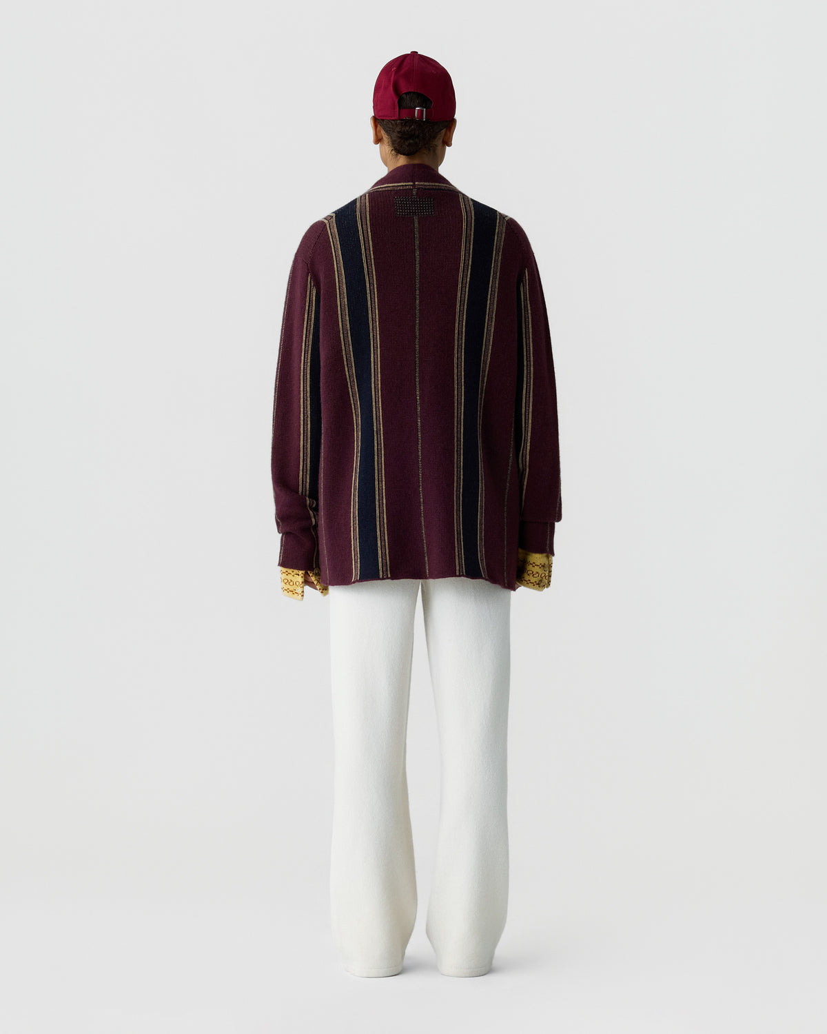 Back view of a burgundy striped cashmere cardigan worn with white trousers and a matching burgundy cap against a light studio background
