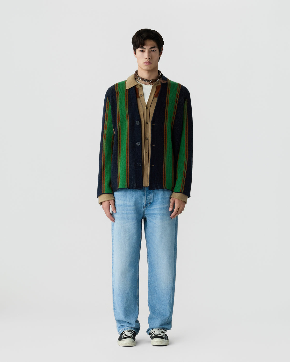 A young man wearing a vertically striped cardigan in green, navy, and brown over a beige shirt, paired with light blue jeans and black-and-white sneakers, standing against a plain white background.