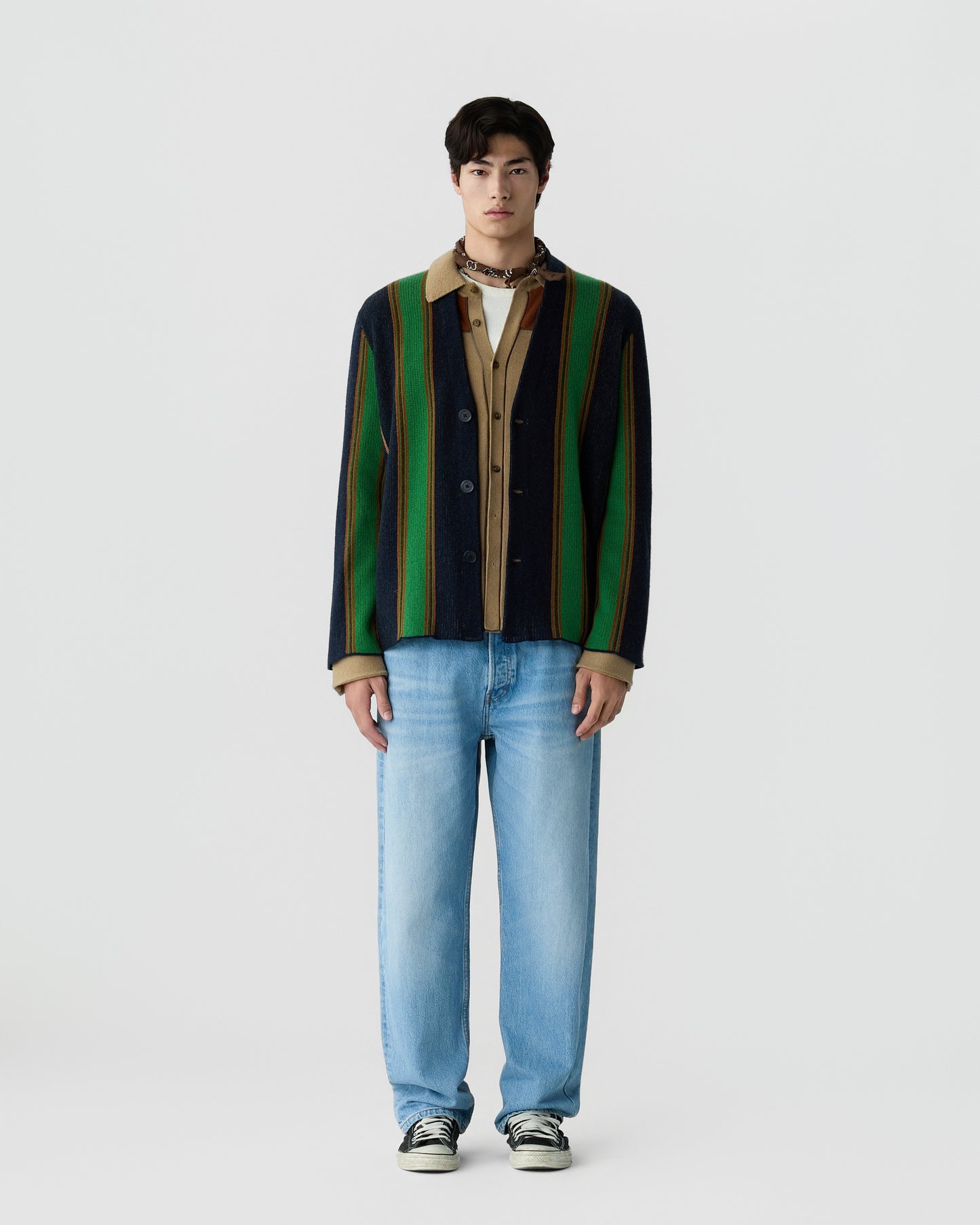 A young man wearing a vertically striped cardigan in green, navy, and brown over a beige shirt, paired with light blue jeans and black-and-white sneakers, standing against a plain white background.