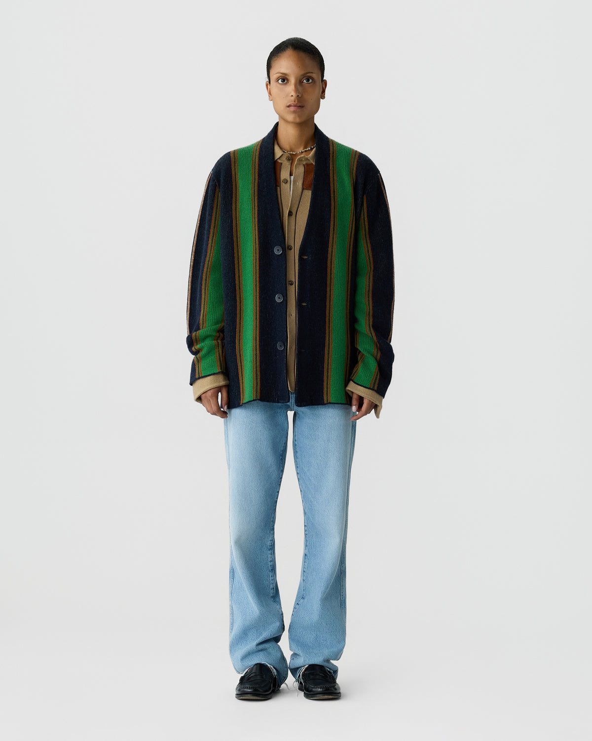 Person wearing a striped cardigan over a collared shirt with light blue jeans and black shoes, standing against a plain light gray background.