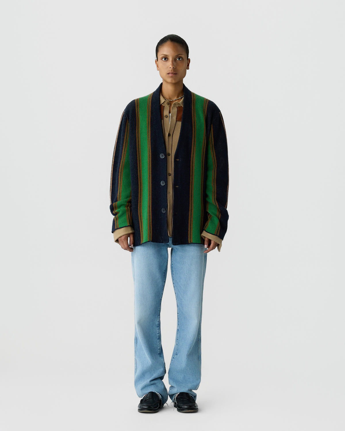 Front view of a person wearing a vertically striped cardigan with green, black, and brown tones over a beige shirt, light blue jeans, and black shoes against a plain white background.