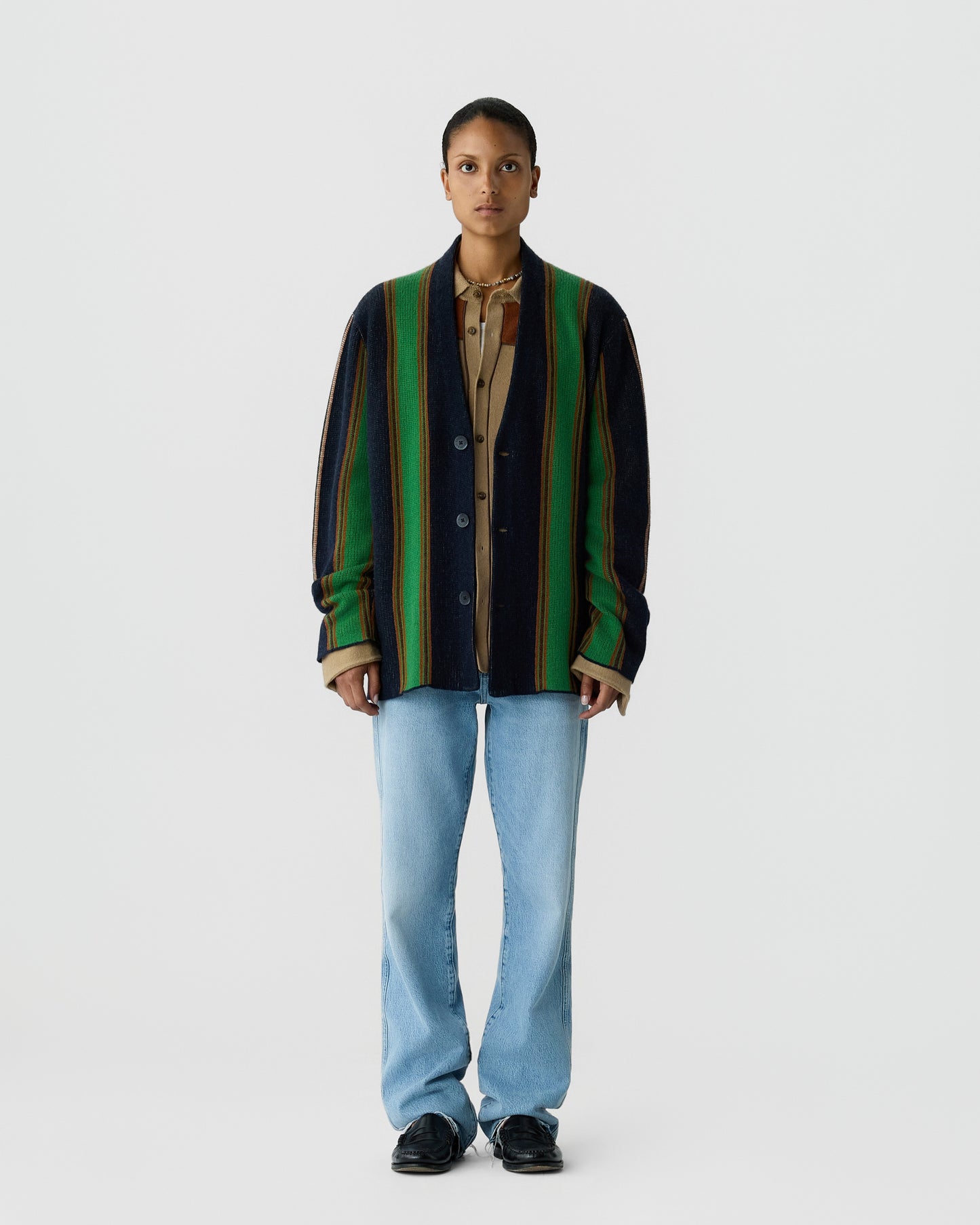 Front view of a person wearing a vertically striped cardigan with green, black, and brown tones over a beige shirt, light blue jeans, and black shoes against a plain white background.