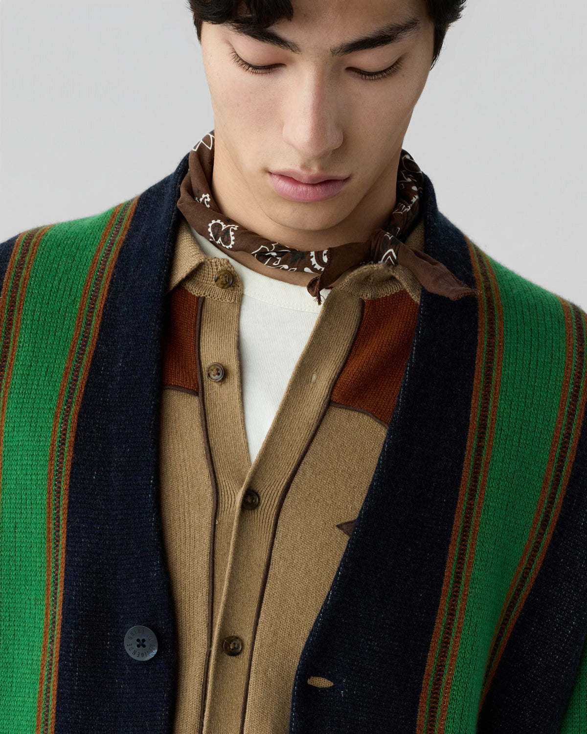 Close-up of a man wearing a brown patterned neck scarf, a beige and brown buttoned cardigan, and a green and navy striped outer garment, looking down with a neutral expression.