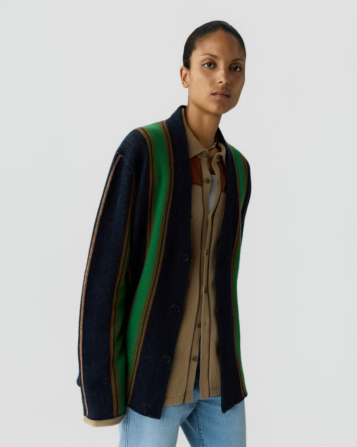 Model wearing a dark cardigan with green and brown stripes over a tan collared shirt and light blue jeans against a plain gray background
