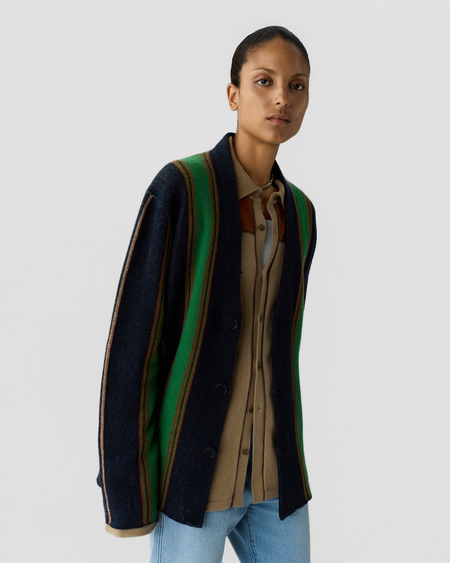 Young woman wearing a navy blue and green striped cardigan over a beige button-up shirt with light blue jeans, standing against a plain gray background.