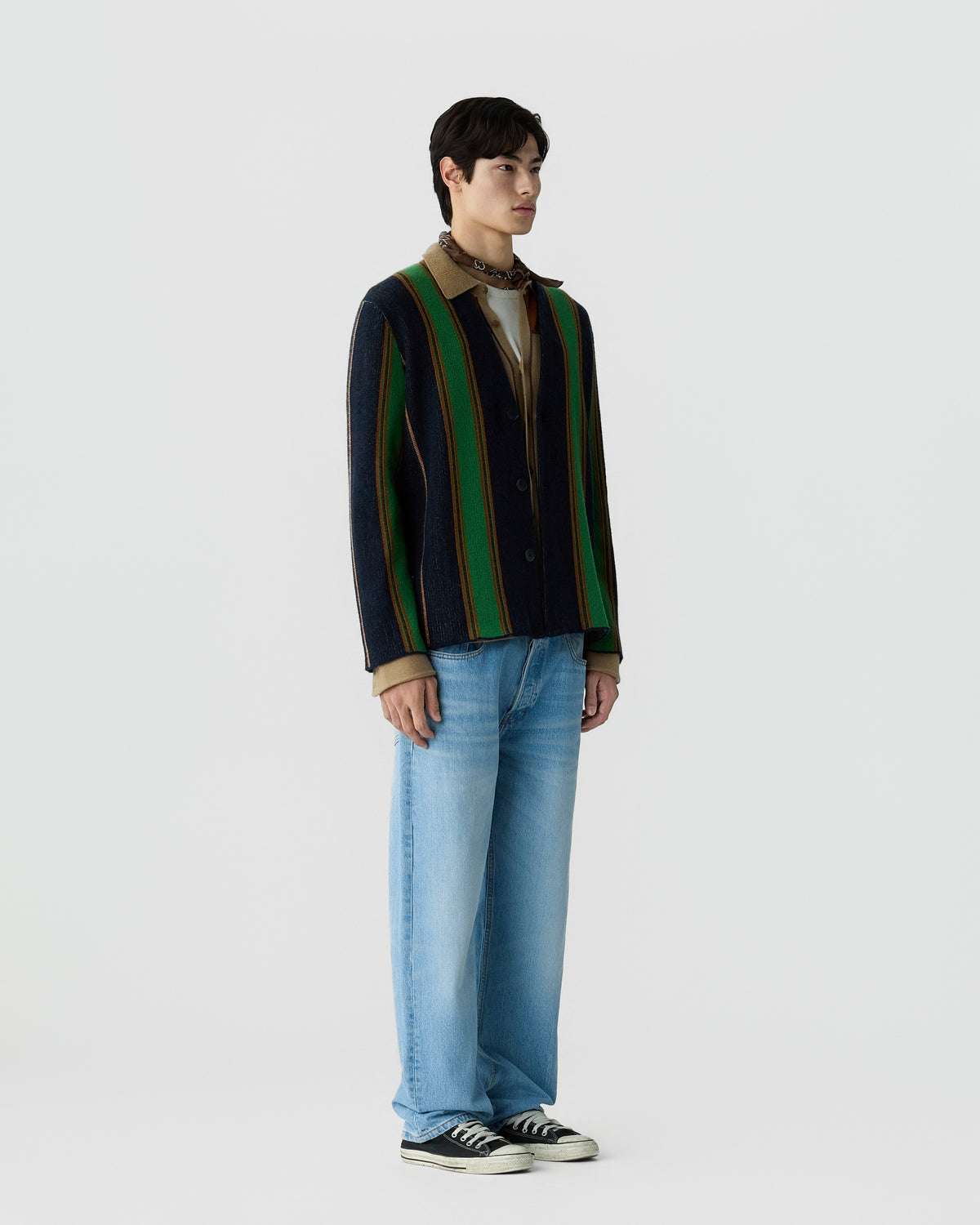 A man stands against a plain background wearing a vertically striped jacket with green, navy, and brown tones over a beige shirt, light blue jeans, and black sneakers.
