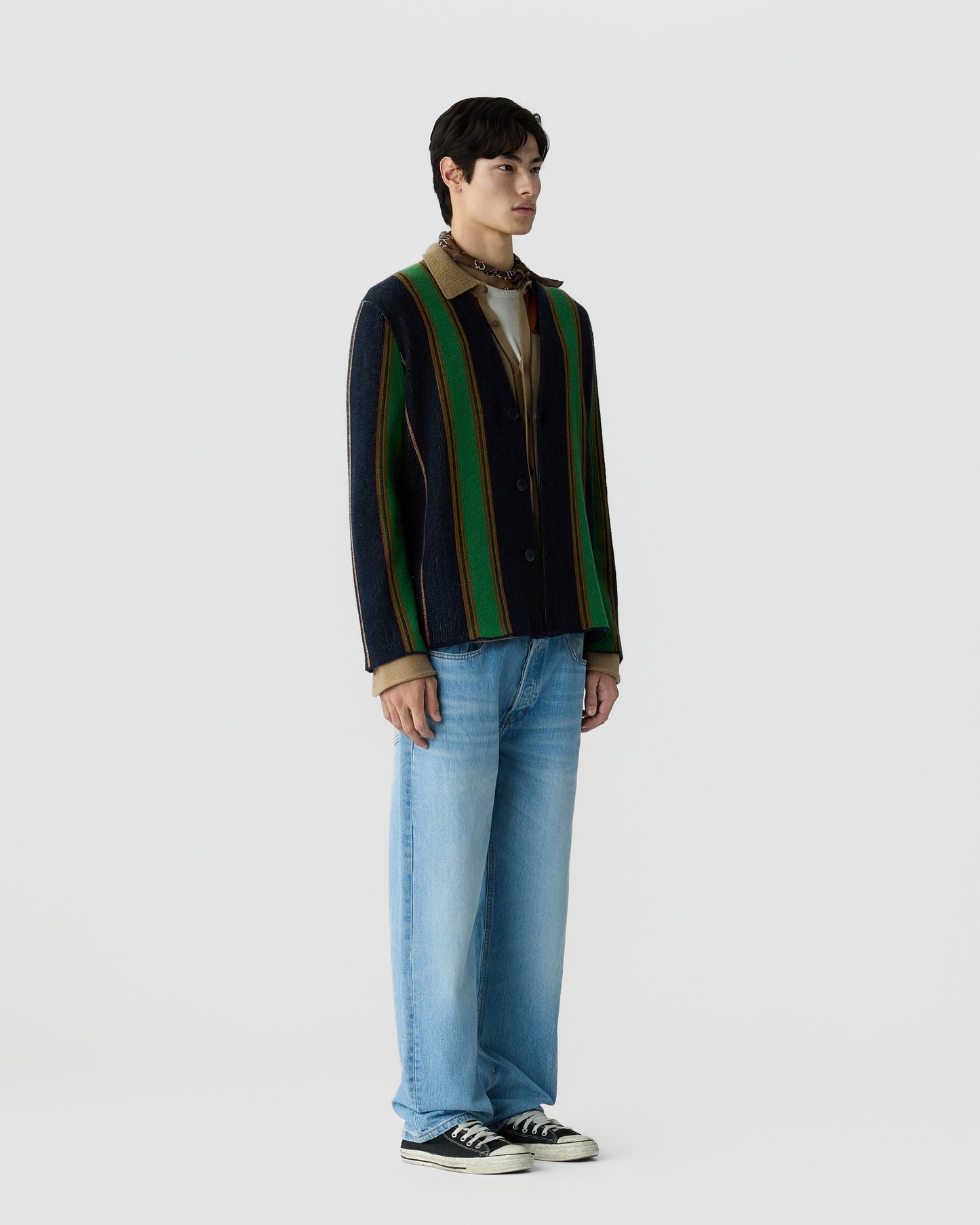 A man stands against a plain background wearing a vertically striped jacket with green, navy, and brown tones over a beige shirt, light blue jeans, and black sneakers.