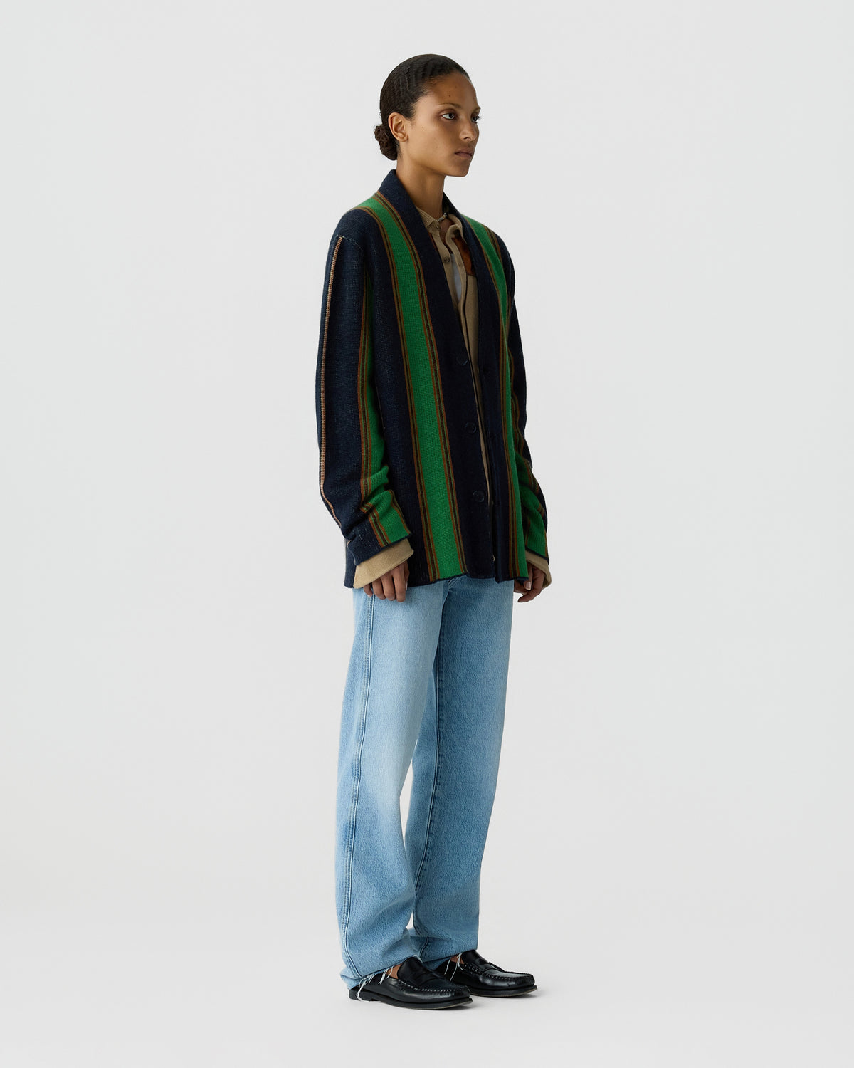 Person standing against a light gray background, wearing a dark green and navy striped sweater, light blue jeans, and black shoes.