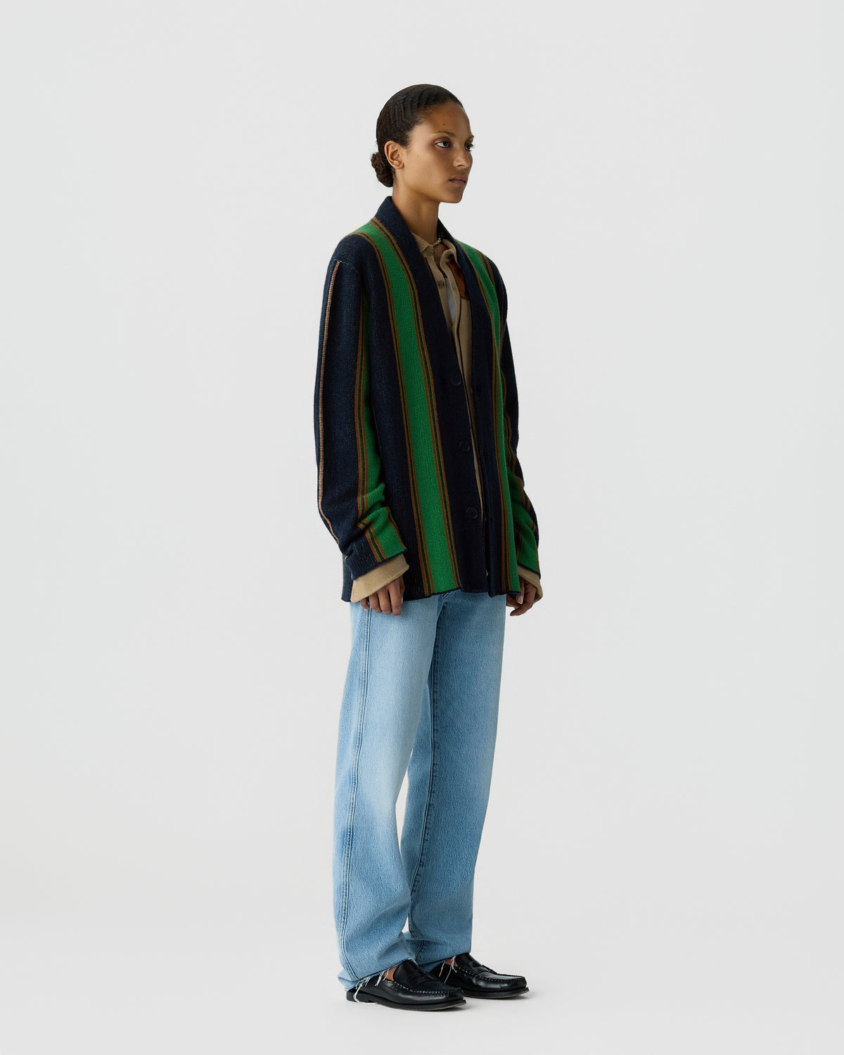 Woman standing against plain background wearing a navy, green, and brown striped cardigan over a beige shirt, light blue jeans, and black shoes in a side profile pose.