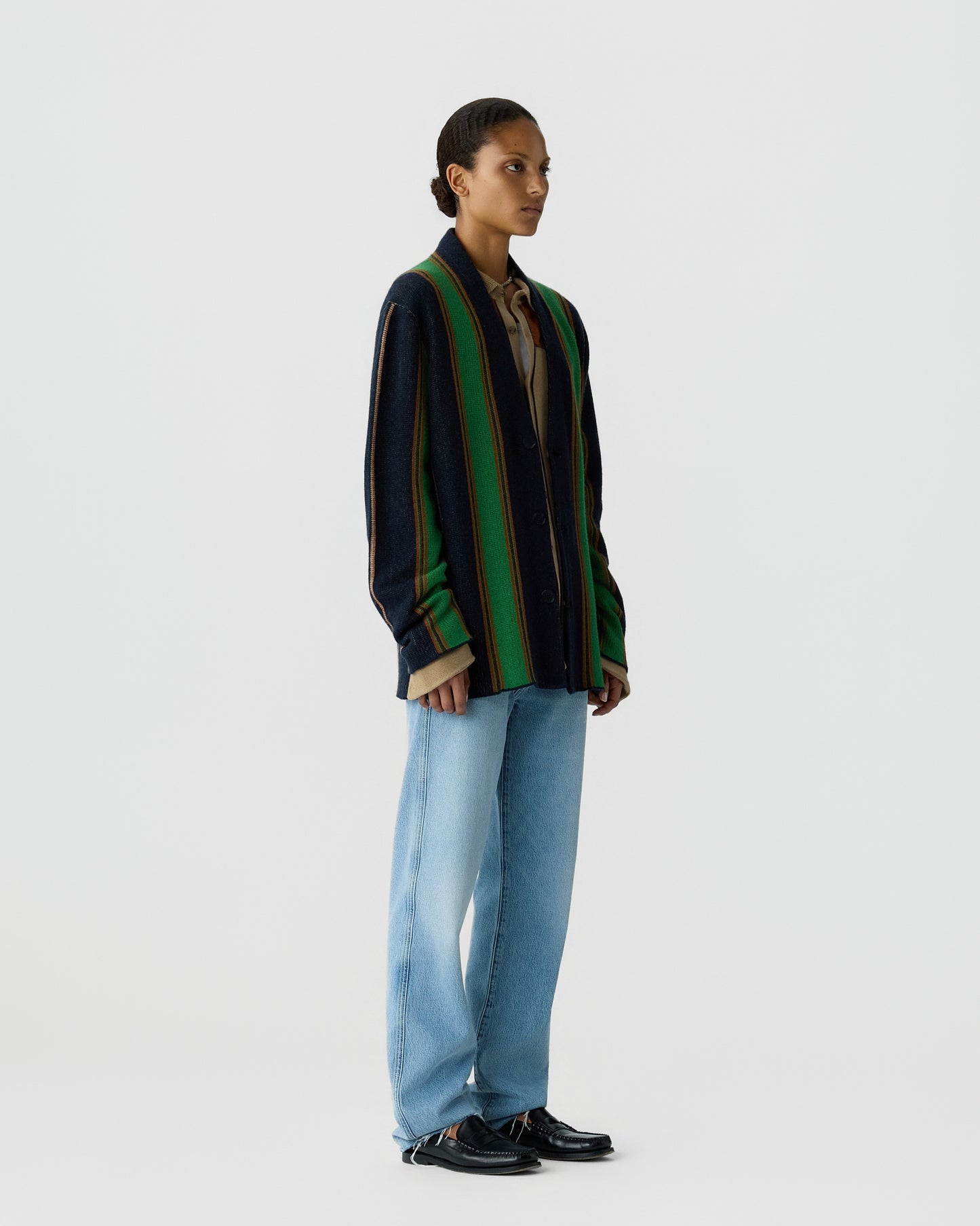 Woman standing against plain background wearing a navy, green, and brown striped cardigan over a beige shirt, light blue jeans, and black shoes in a side profile pose.