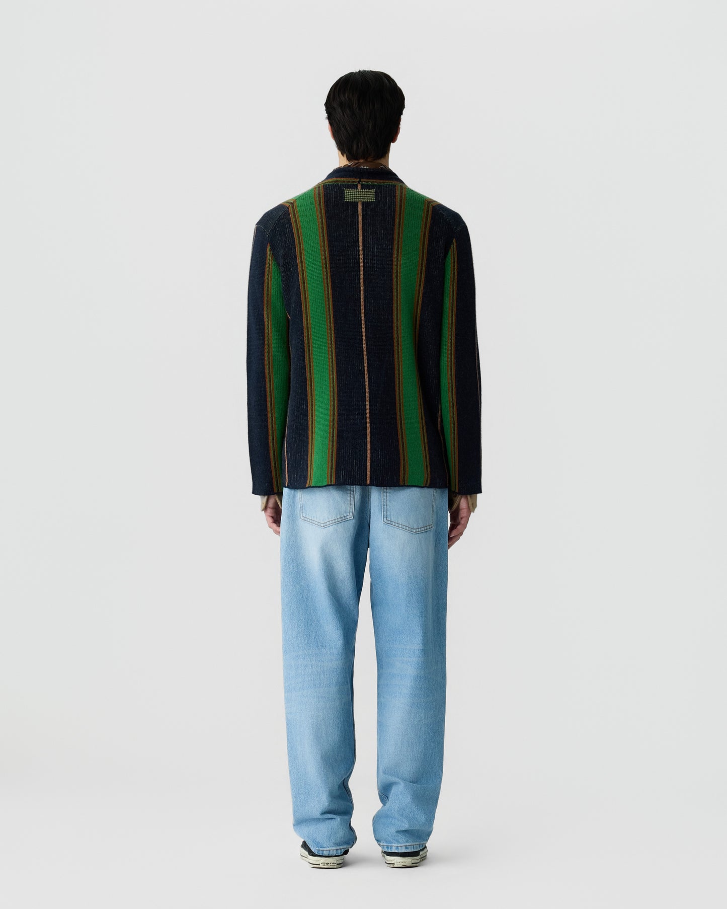 Man wearing a dark jacket with vertical green and yellow stripes on the back paired with light blue jeans and sneakers, standing against a plain white background.
