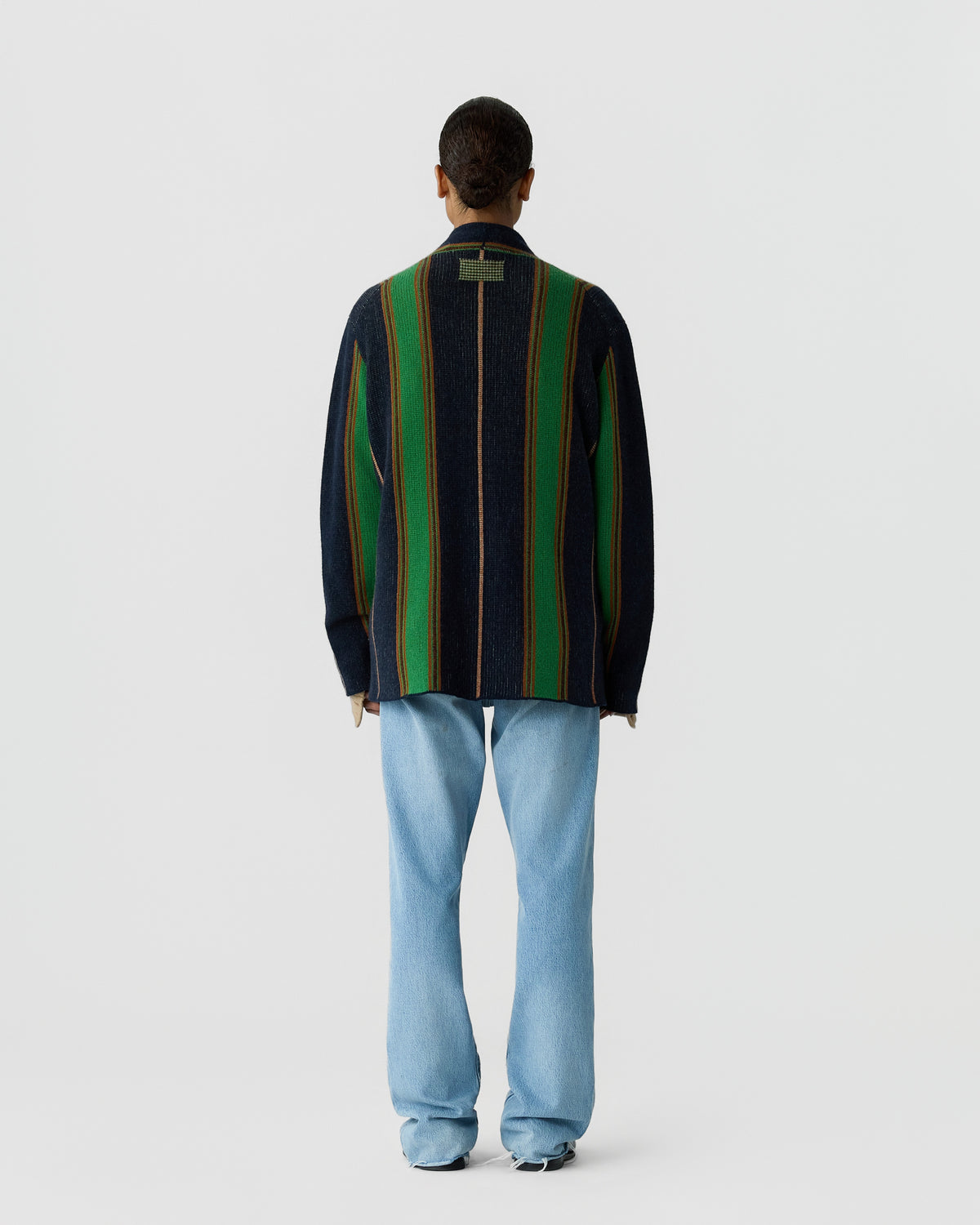 Person standing with their back to the camera wearing a dark sweater with vertical green and orange stripes and light blue jeans against a plain white background.