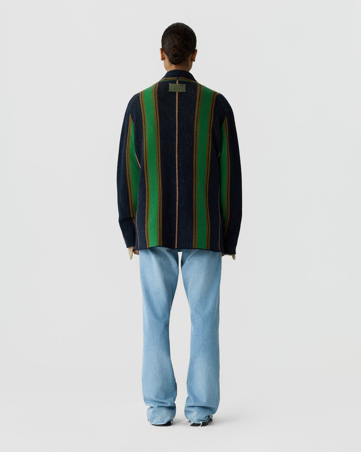 Person seen from behind wearing a dark striped knit cardigan with green and brown accents, light blue jeans, and dark shoes against a gray background
