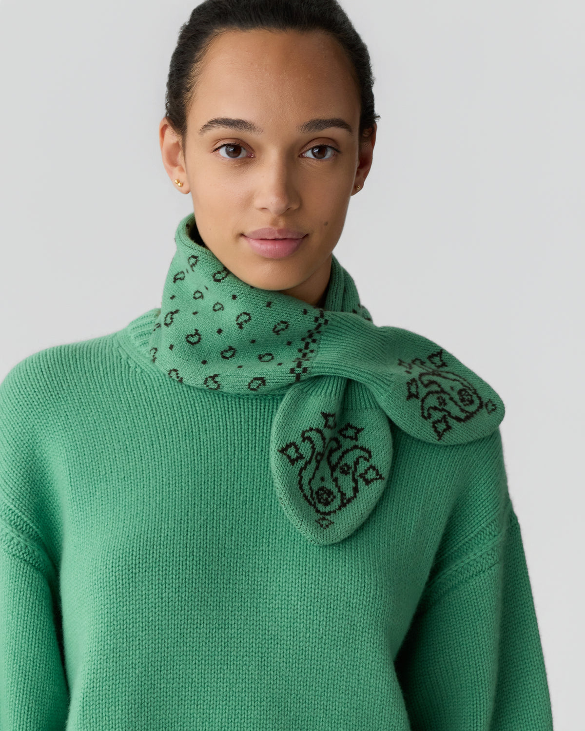 Young woman wearing a green knitted sweater and a matching green scarf with a black paisley and dot pattern tied around her neck against a plain light background.
