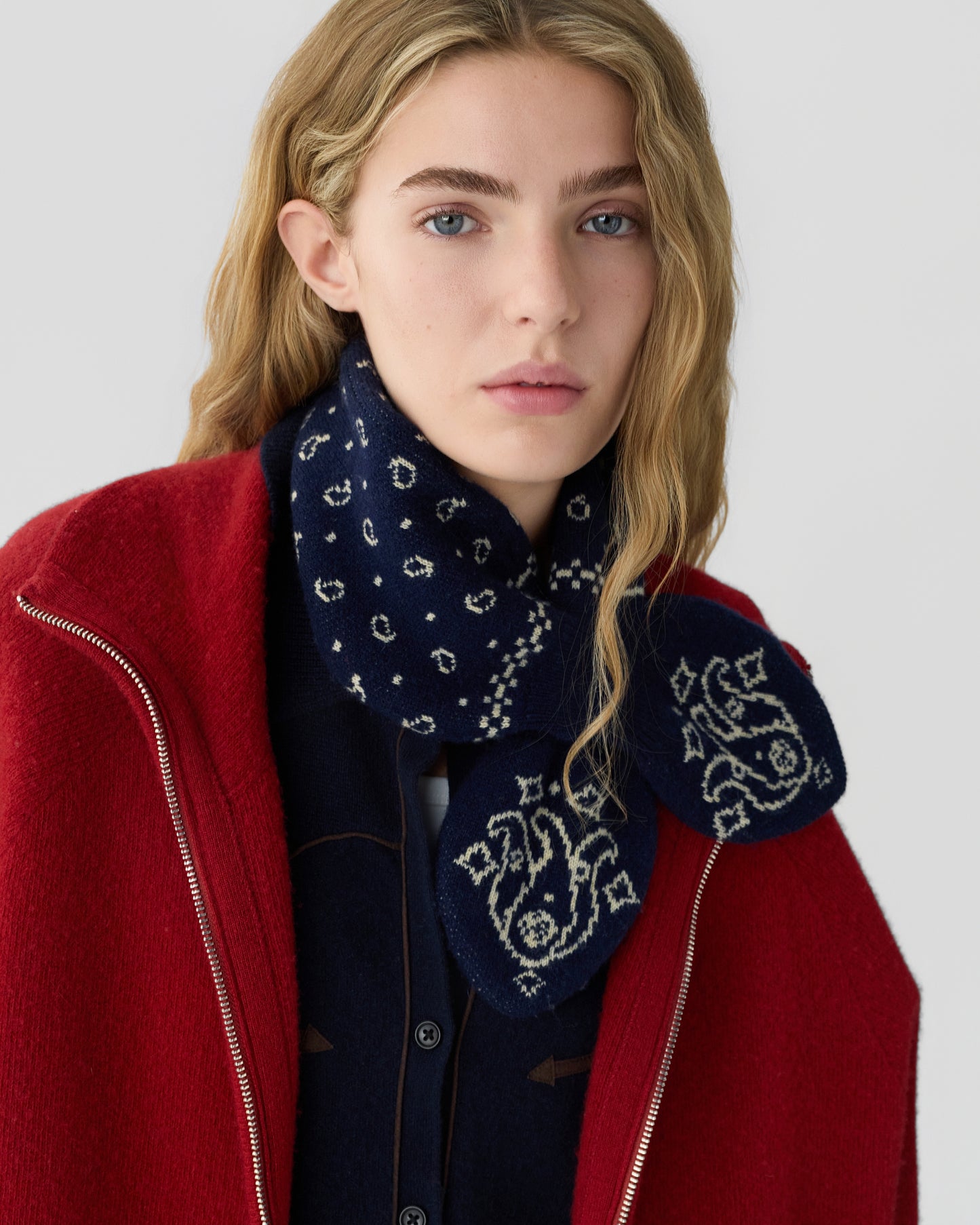 Young woman with wavy blonde hair wearing a dark blue patterned cashmere scarf and a red zip-up jacket against a neutral background.
