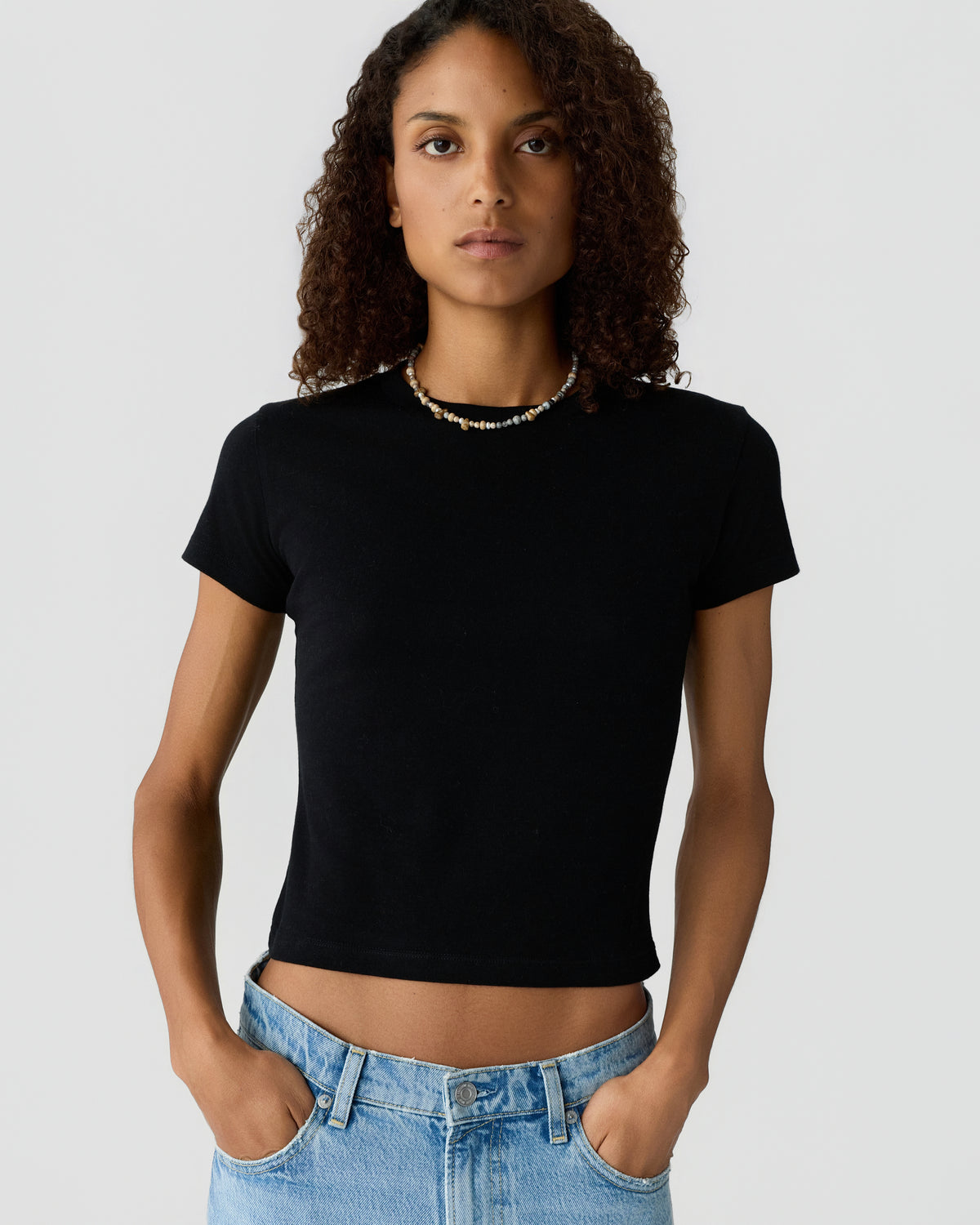 Woman with curly hair wearing a fitted black short sleeve top and light blue jeans, standing against a plain light gray background with hands in pockets.