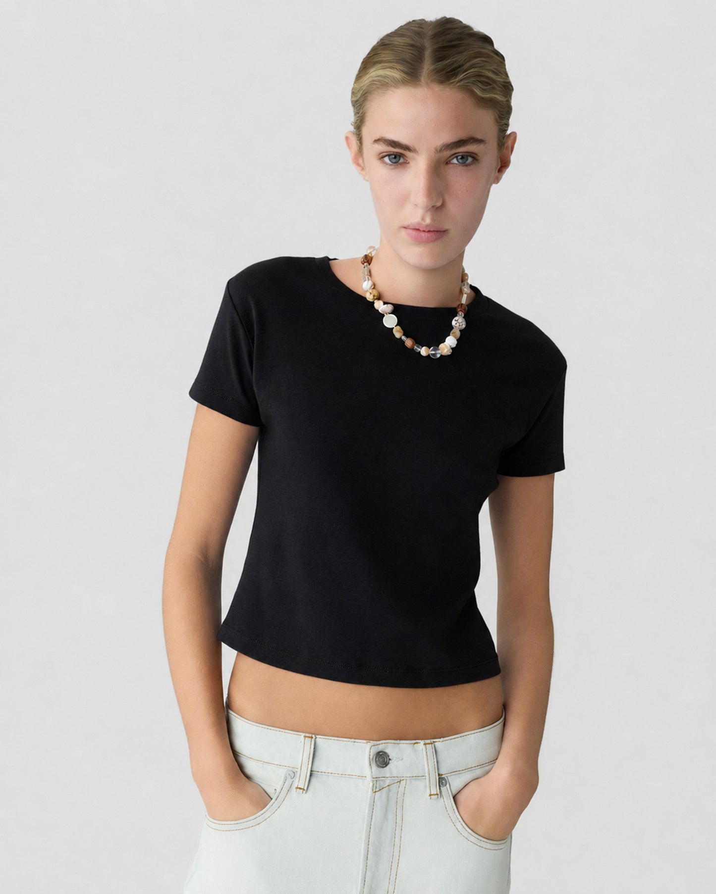 Woman wearing a fitted black short sleeve shirt with light wash jeans and a beaded necklace, standing against a light gray background with hands in pockets