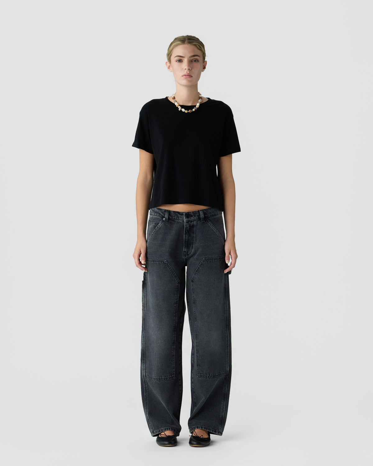 Full-length portrait of a woman wearing a black short-sleeve top, loose dark gray jeans, black shoes, and a necklace against a light gray background.