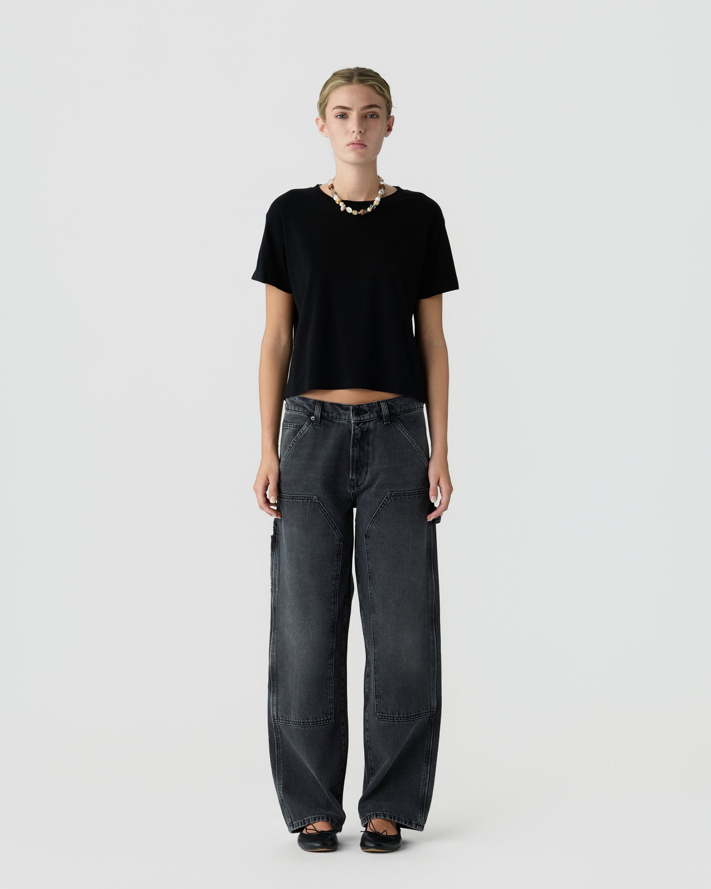 Full-length portrait of a woman wearing a black short-sleeve top, loose dark gray jeans, black shoes, and a necklace against a light gray background.