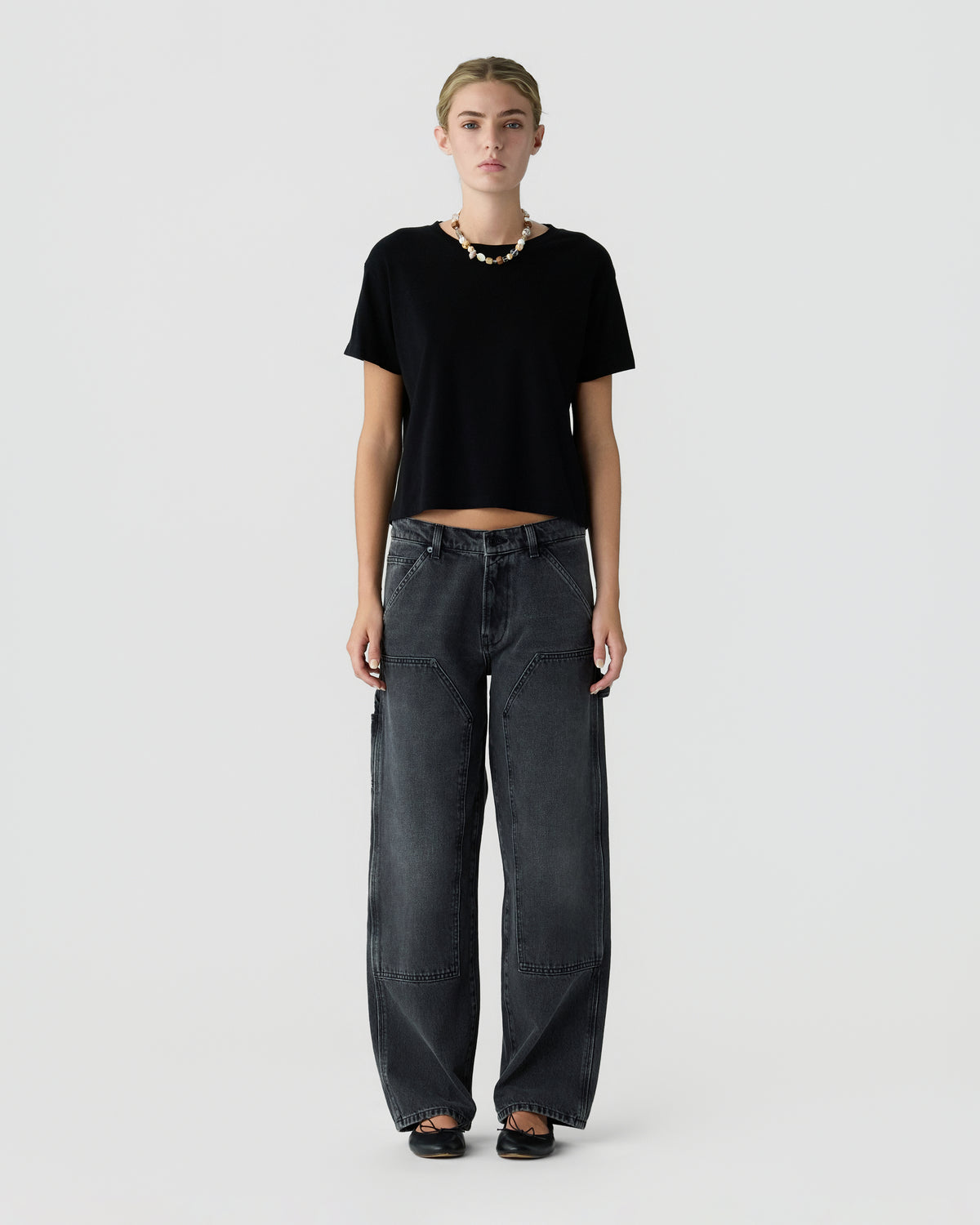 Model wearing a black cropped T shirt, dark gray loose fit jeans, black shoes, and a beaded necklace against a light gray studio background
