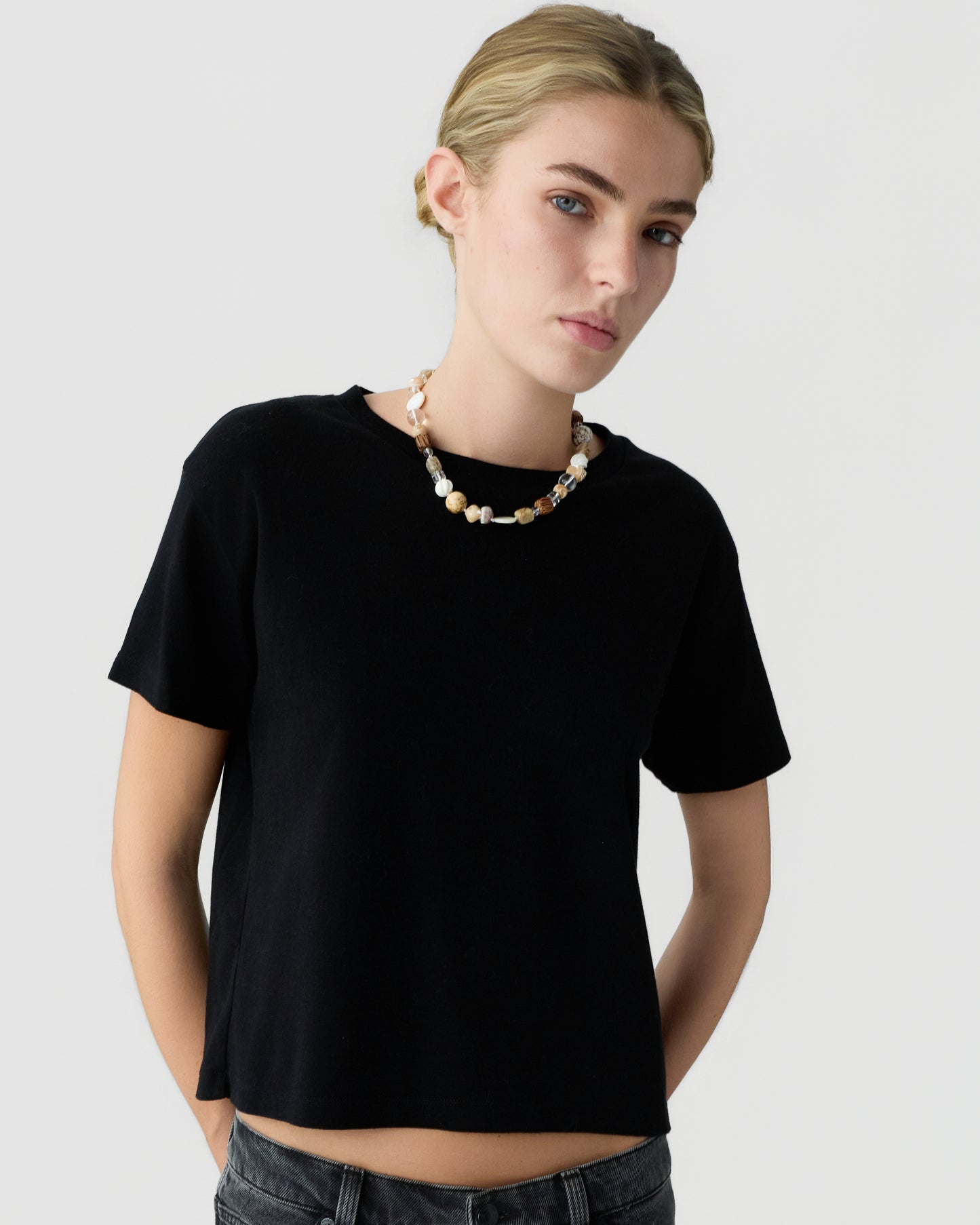 Young woman with blonde hair wearing a plain black short sleeve shirt and beaded necklace against a light gray background