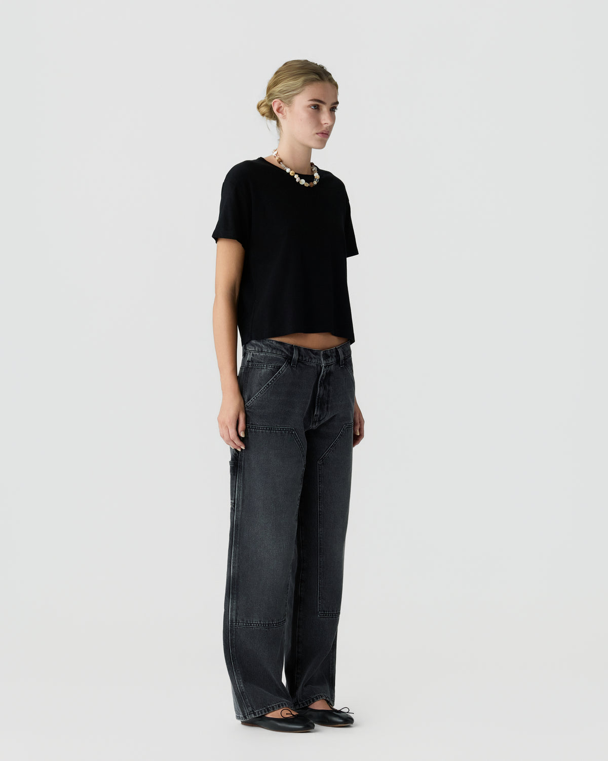 Full-length view of a woman wearing a black short-sleeve top with dark wide-leg jeans and flat shoes against a light gray background.