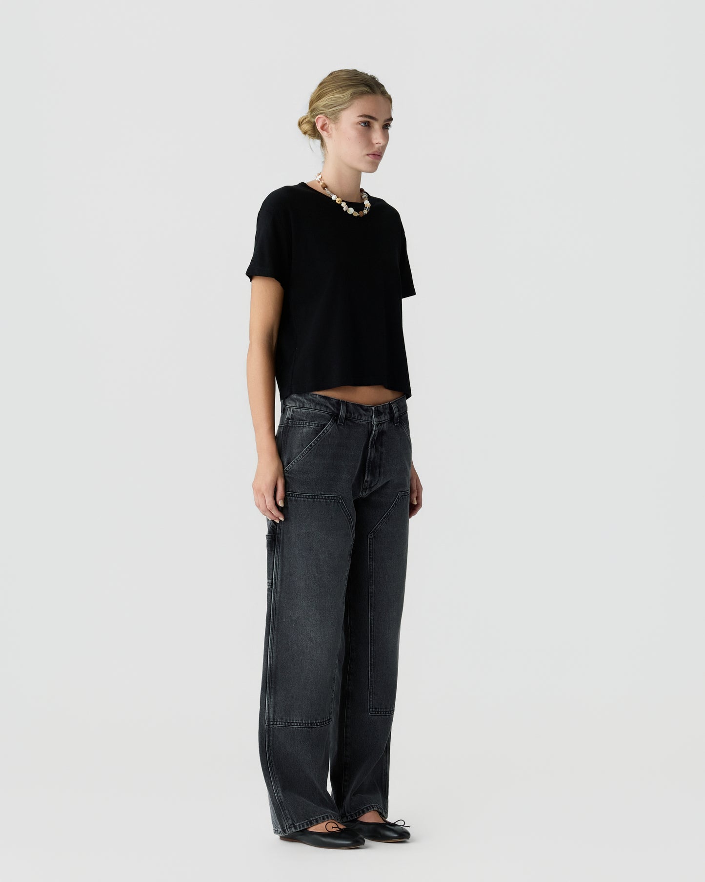Full-length view of a woman wearing a black short-sleeve top with dark wide-leg jeans and flat shoes against a light gray background.