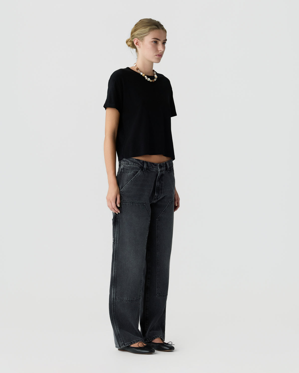 Model wearing a black short sleeve top, dark wide leg jeans, black shoes, and a chunky necklace against a light gray studio background