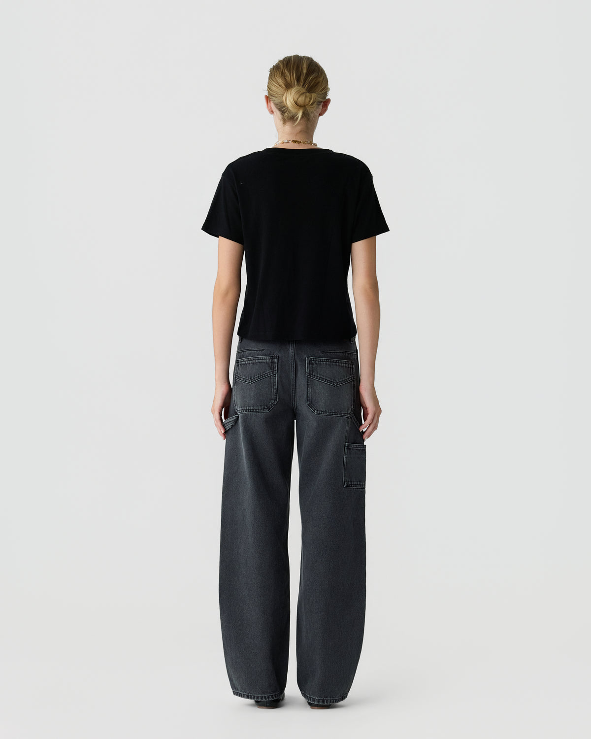 Back view of a person with blonde hair wearing a black short sleeve shirt and dark gray wide leg utility jeans against a light gray background.