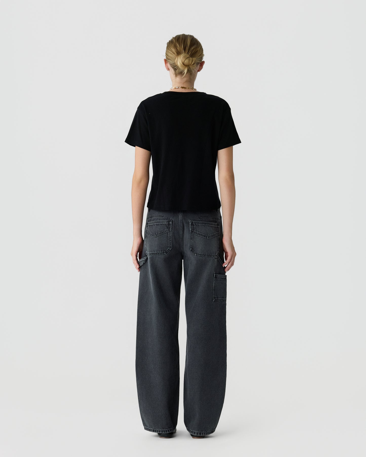 Back view of a person with blonde hair wearing a black short sleeve shirt and dark gray wide leg utility jeans against a light gray background.