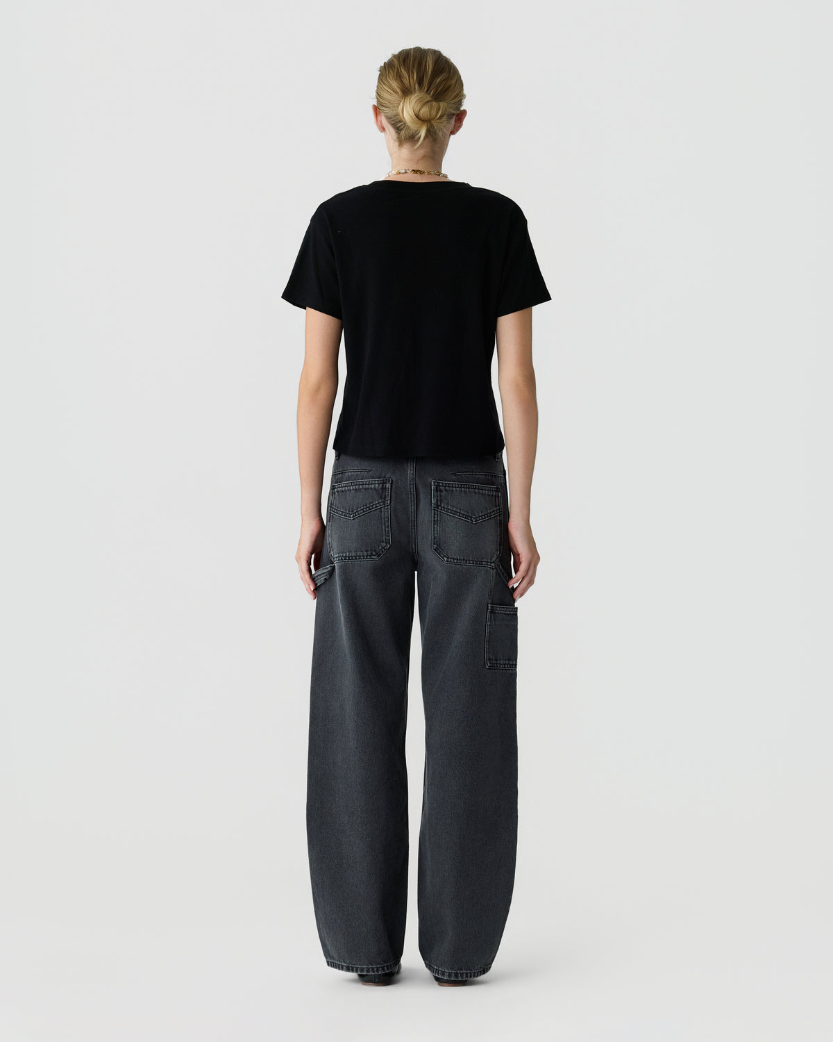 Back view of a model wearing a black short sleeve T shirt and relaxed dark gray wide leg jeans with utility pockets against a light gray background