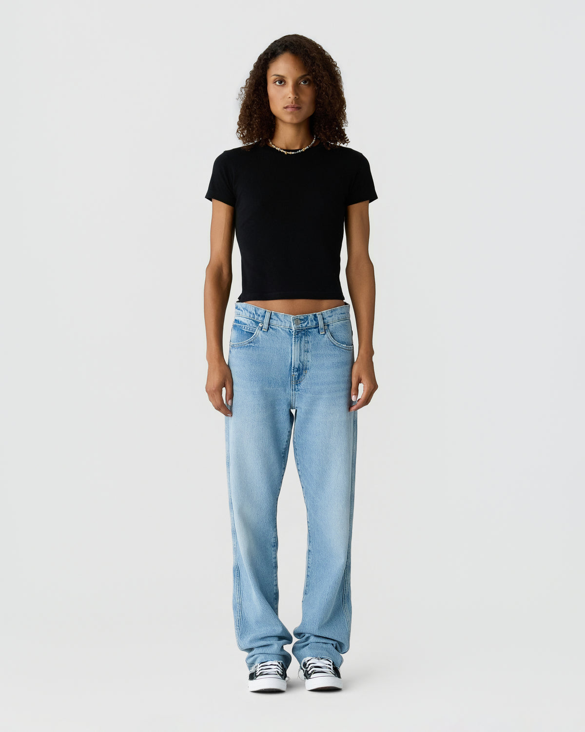 Woman standing against a light gray background wearing a black cropped T shirt light blue relaxed jeans and white sneakers