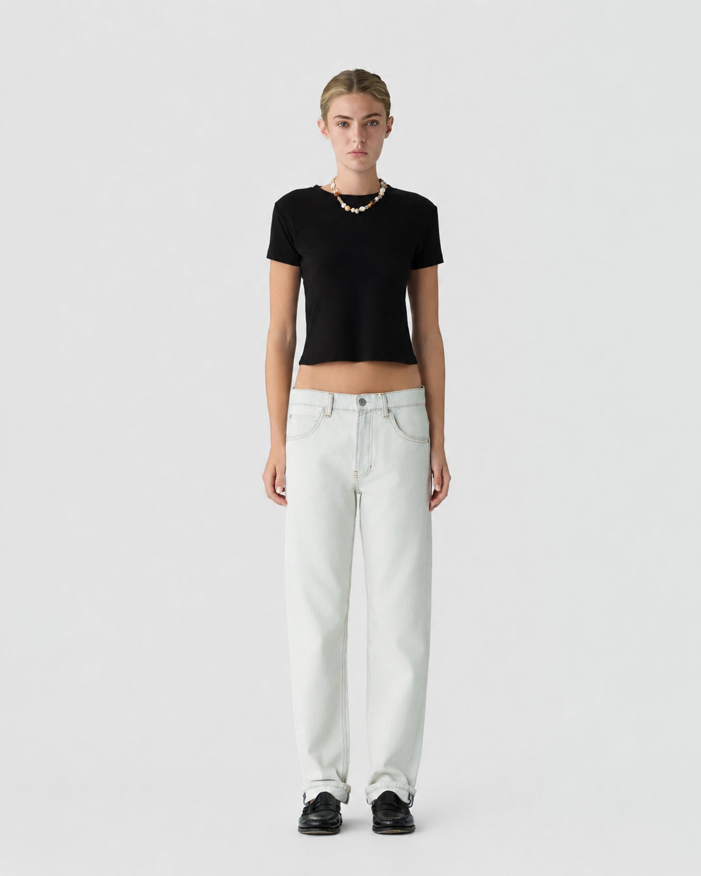 Model wearing a black short sleeve cropped top with white wide leg jeans and black shoes against a light gray studio background